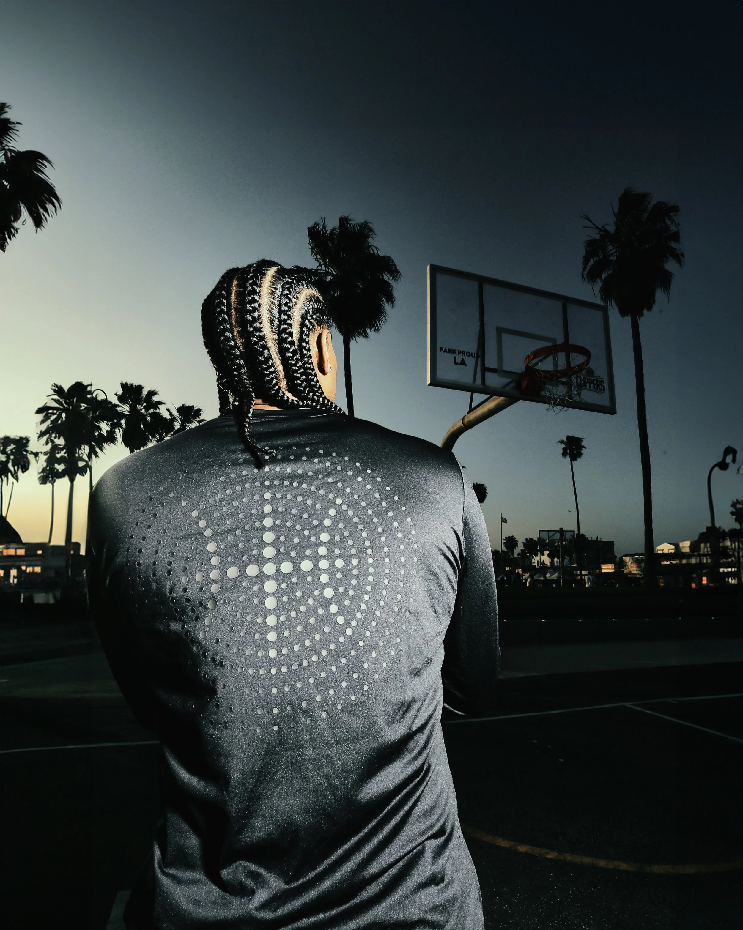 A person with braided hair and a sleek sports jacket standing in a dark outdoor basketball court, with a basketball hoop and some palm trees visible in the background during dusk.