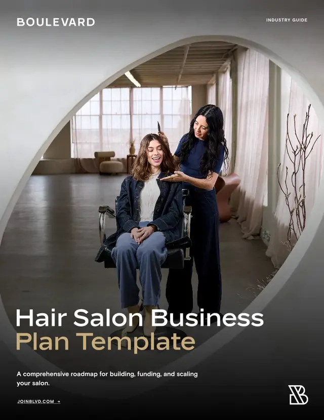 A woman in a blue shirt giving a haircut to a seated woman in a hair salon. The salon has large windows with curtains and an open, modern interior design.
