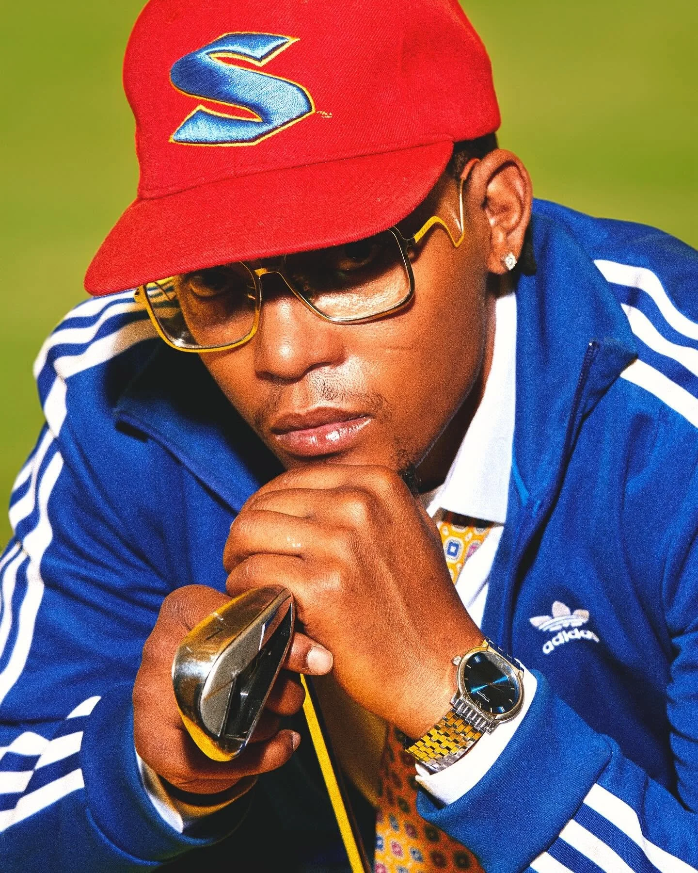 A man wearing a red cap with a large blue 'S' logo, glasses, a blue Adidas jacket, a gold watch, and earrings, holding a silver device against his lips with a green background.