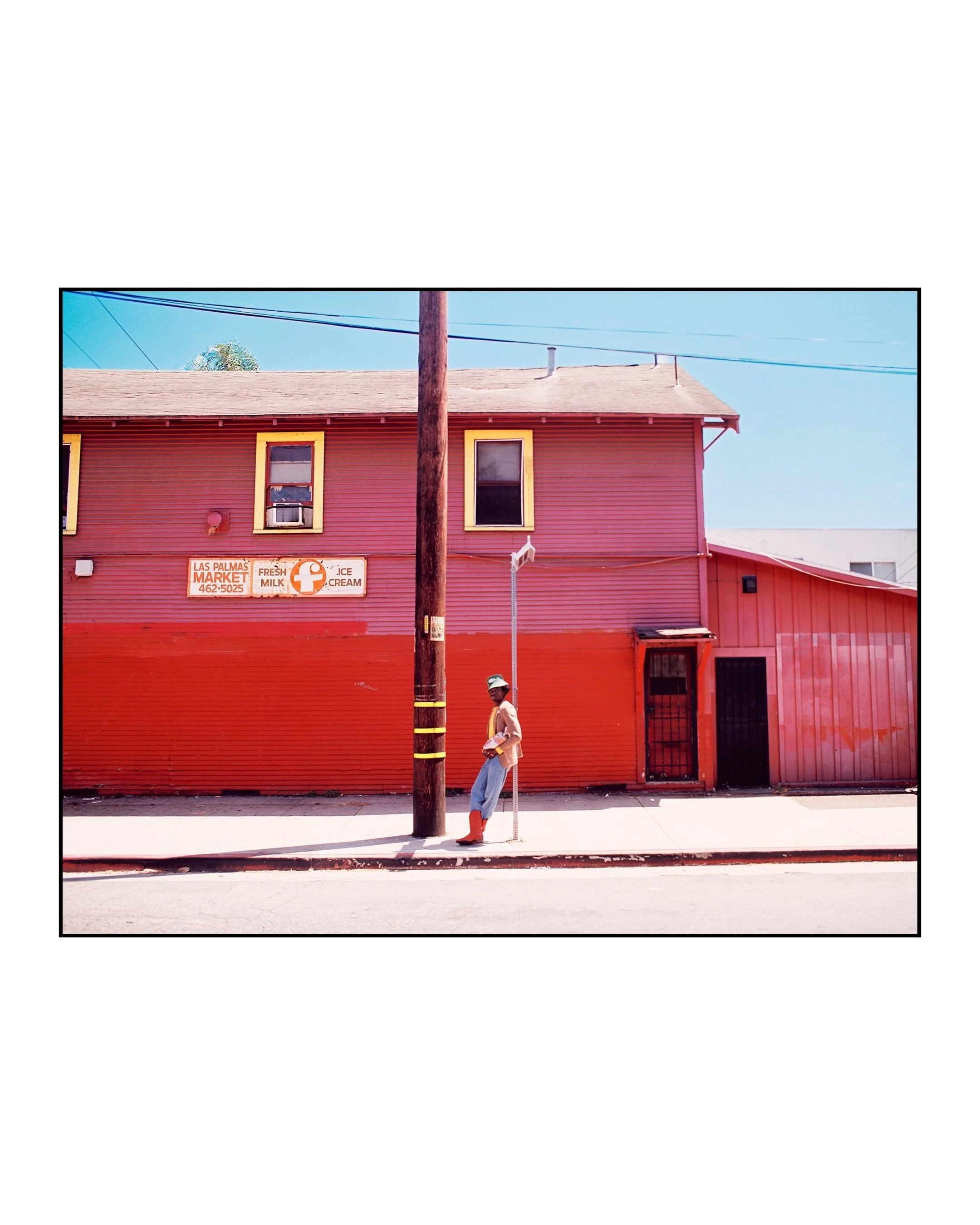 A person in a beige jacket, blue jeans, orange boots, and a wide-brimmed hat standing on the sidewalk beside a red building and a utility pole in an urban area. The building has yellow window trim, a sign advertising milk, ice cream, and a phone number, and a small entrance door.