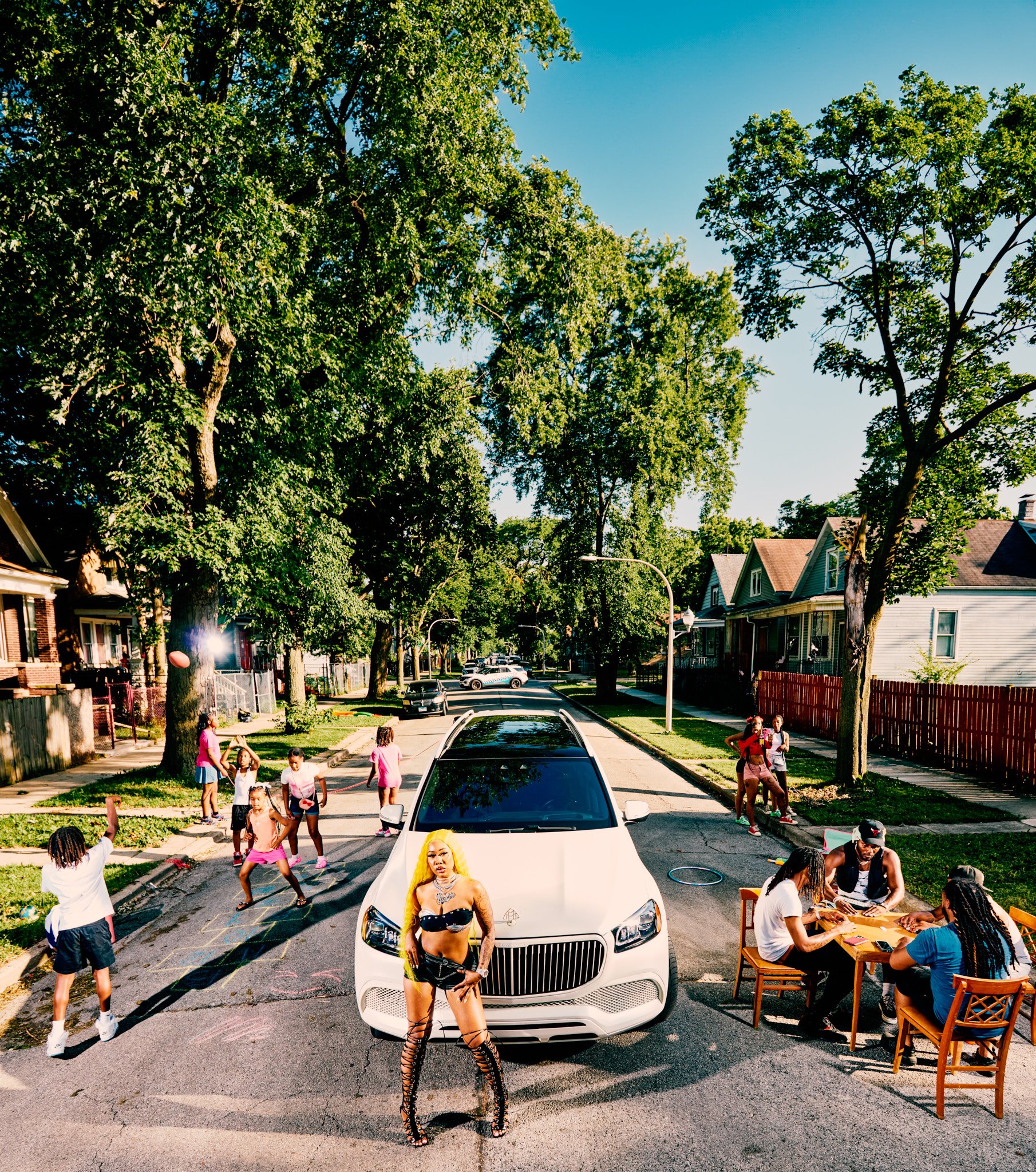 A street scene with children playing, people sitting at a table, a white luxury car parked in the middle, and a woman with long yellow hair and a black outfit standing in front of the car. The background shows trees, houses, and a clear blue sky.