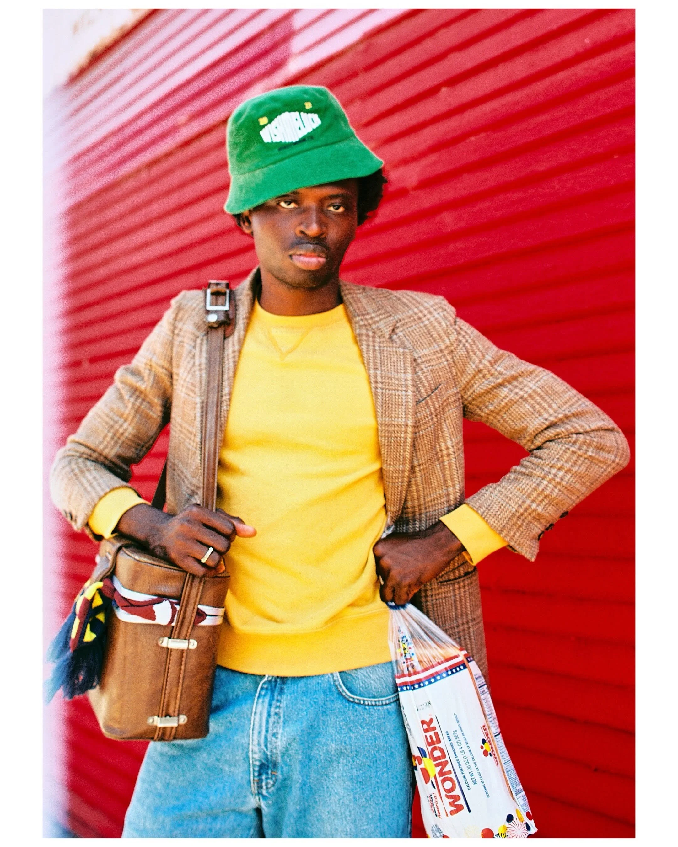 A person wearing a green bucket hat, a yellow shirt, a plaid blazer, blue jeans, and carrying a brown shoulder bag standing in front of a red corrugated metal wall, holding fireworks box.