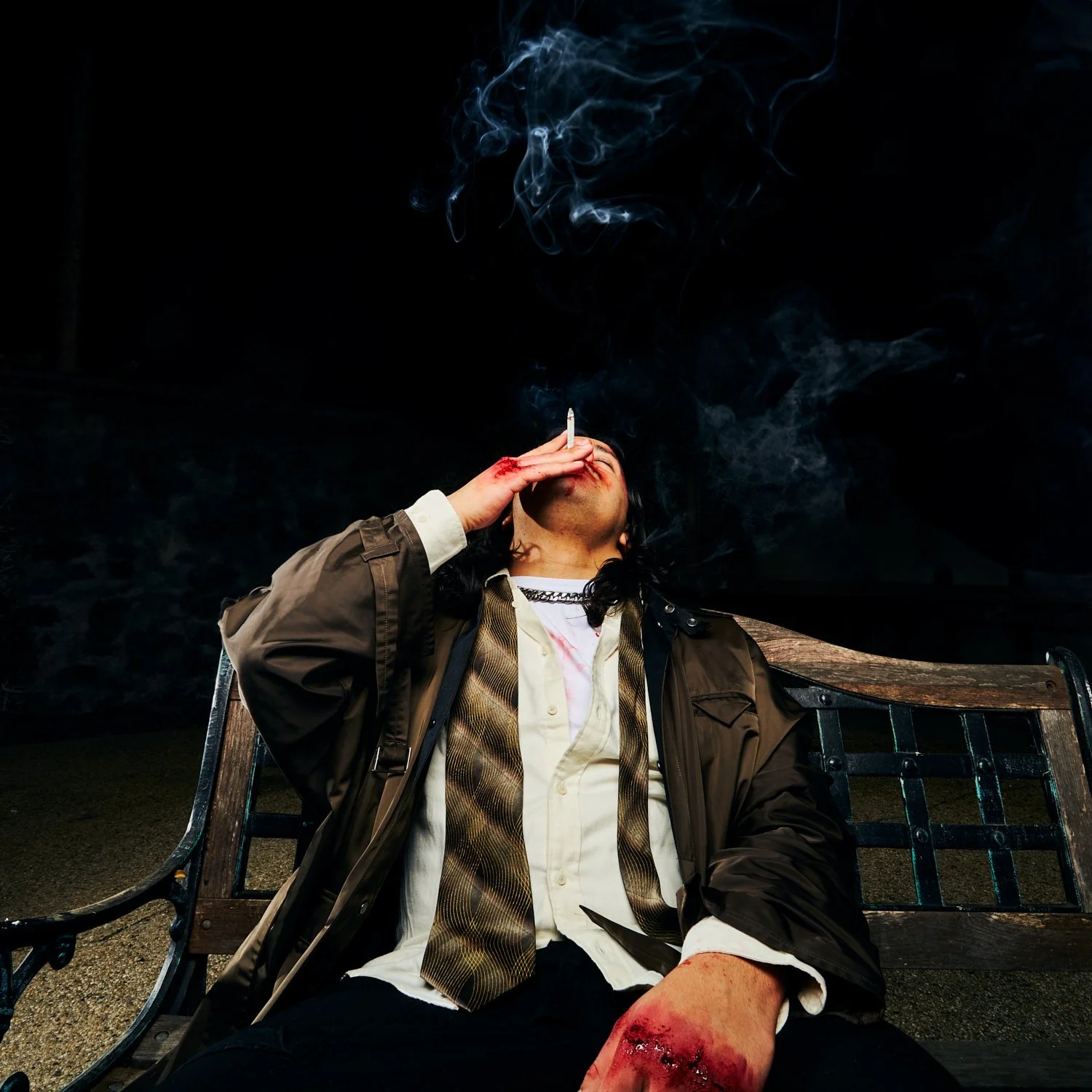 A man sitting on a park bench at night, with a cigarette in his mouth and a bloody injury on his leg, appears intoxicated or exhausted, with dark background and smoke rising from the cigarette.