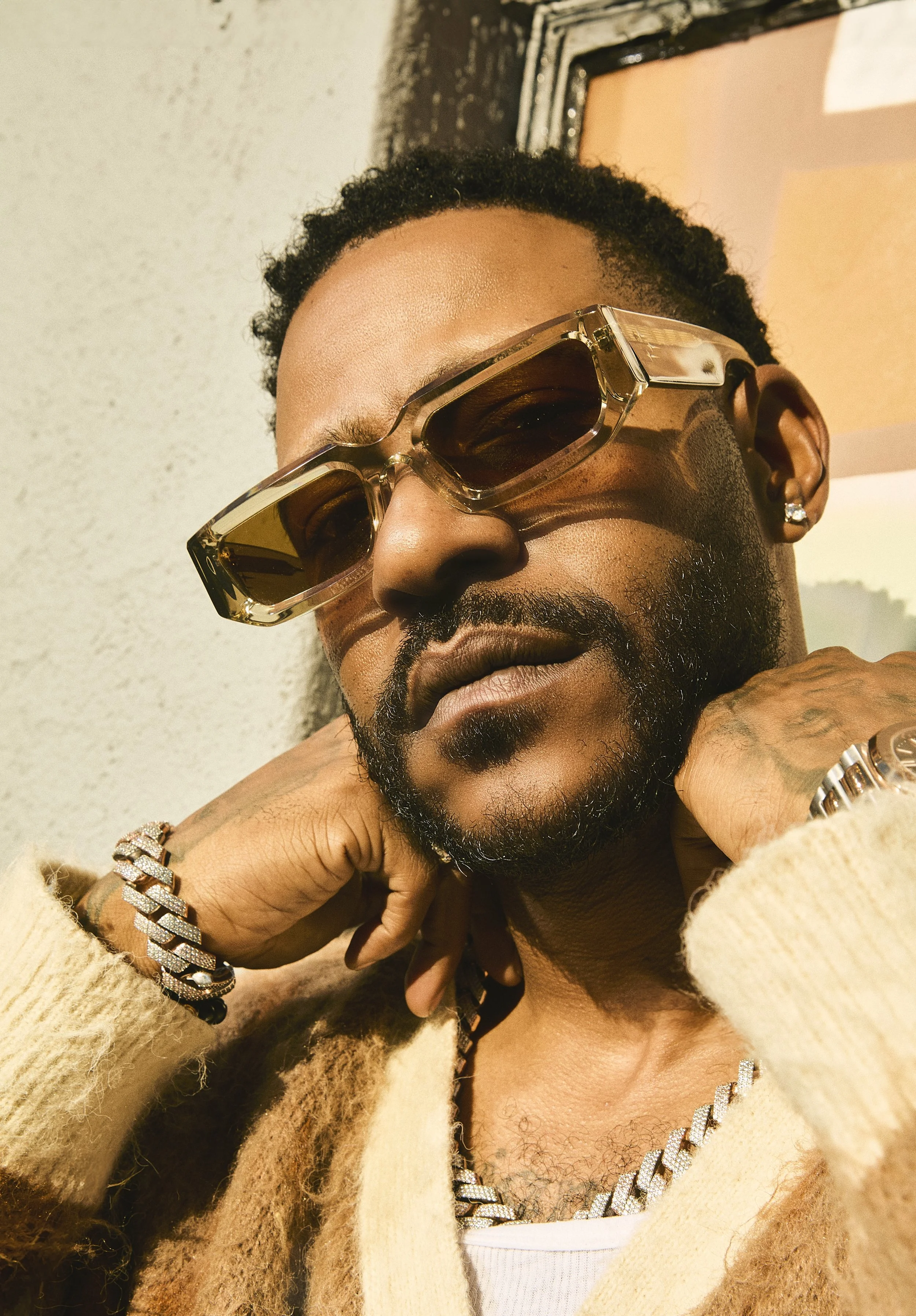 Close-up of a man with a beard wearing sunglasses, a diamond earring, and jewelry, resting his face on his hand, dressed in a cream-colored fur coat and gold chain, against a background with a mirror and textured wall.