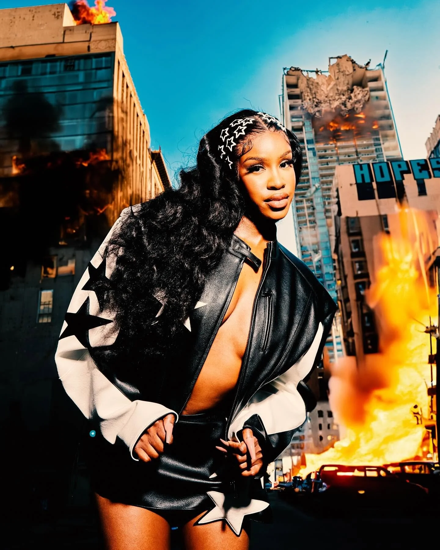 A woman with long black hair wearing a star-patterned headband and a black leather jacket with star designs, standing in front of buildings on fire in an urban setting.