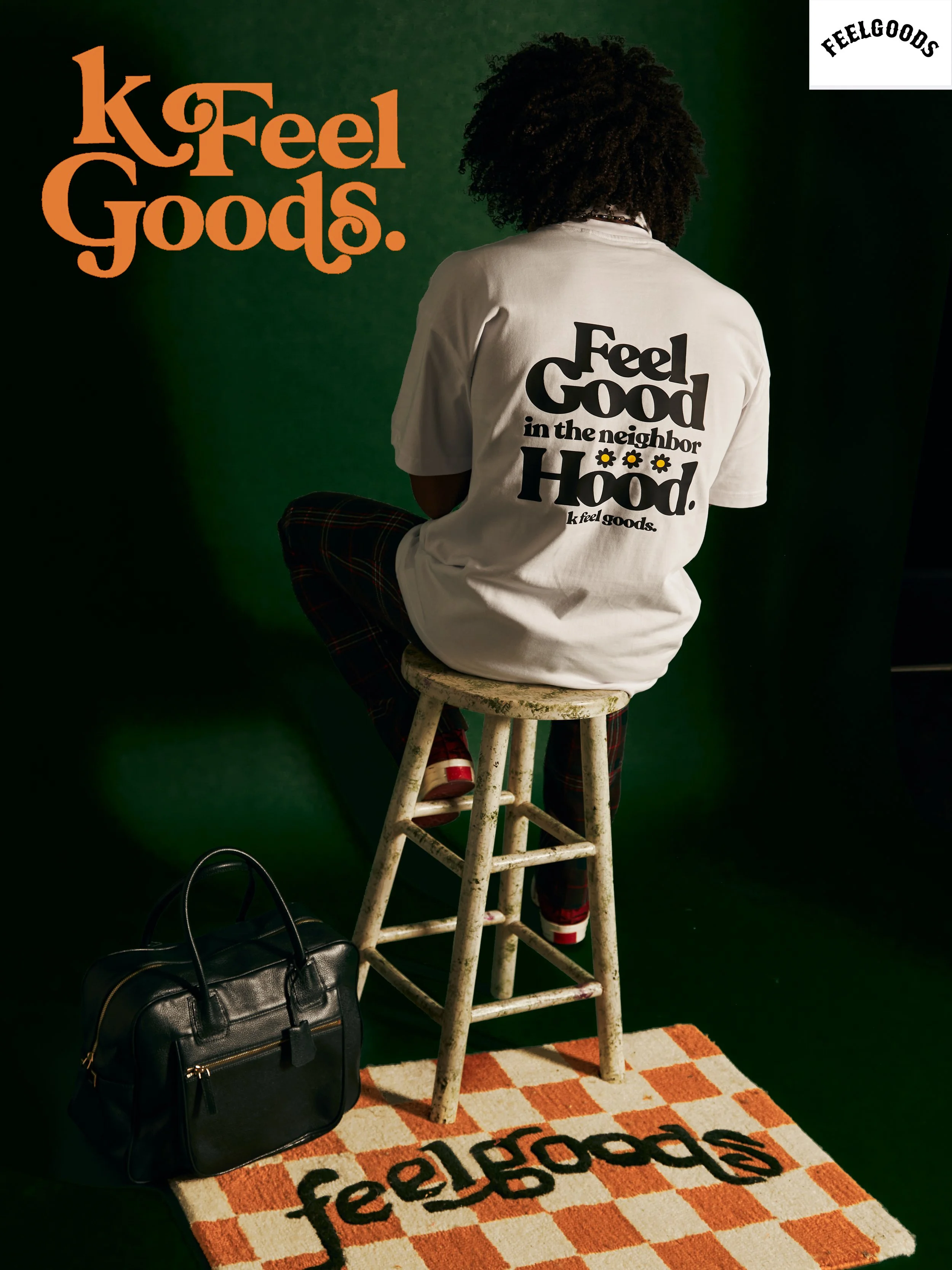 A person sitting on a stool facing away, wearing a white shirt with black text and daisies, plaid pants, and red shoes. There is a black handbag on the floor nearby, an orange and white checkered rug with the word "FeelGood" printed on it, and a green background. The person is facing a wall with the words "K! Feel Goods" written in orange. A white corner in the top right has the Feelsgood logo.