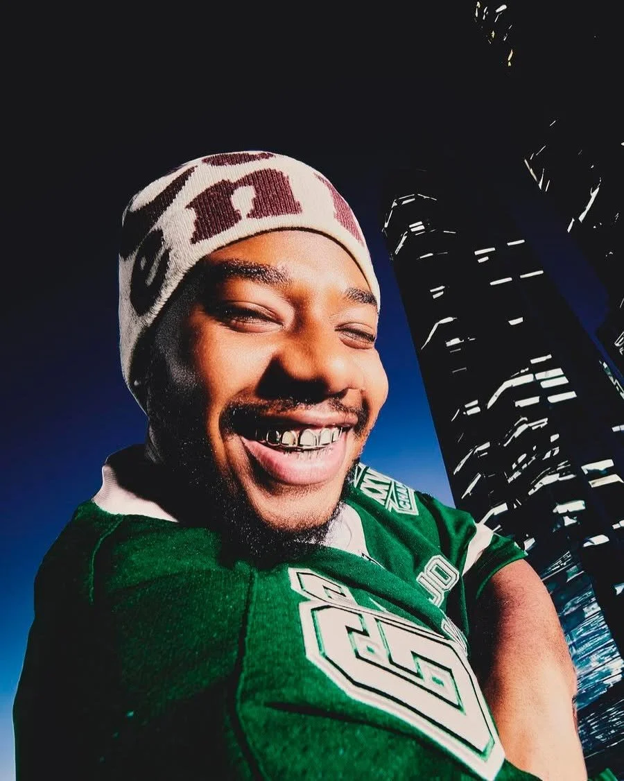 Smiling man wearing a beanie and green sports jersey, standing in front of a tall modern building at night.