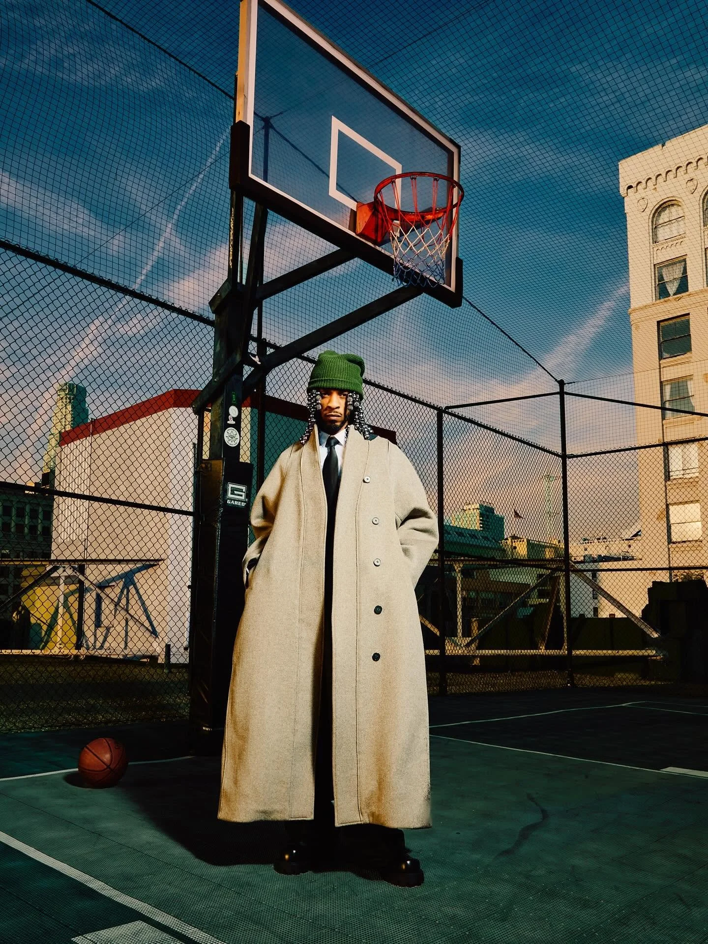 A person with braided hair wearing a green beanie, beige overcoat, white shirt, and black tie standing on an outdoor basketball court with a basketball at their feet, near a basketball hoop with a backboard, enclosed by a chain-link fence in a cityscape during sunset.