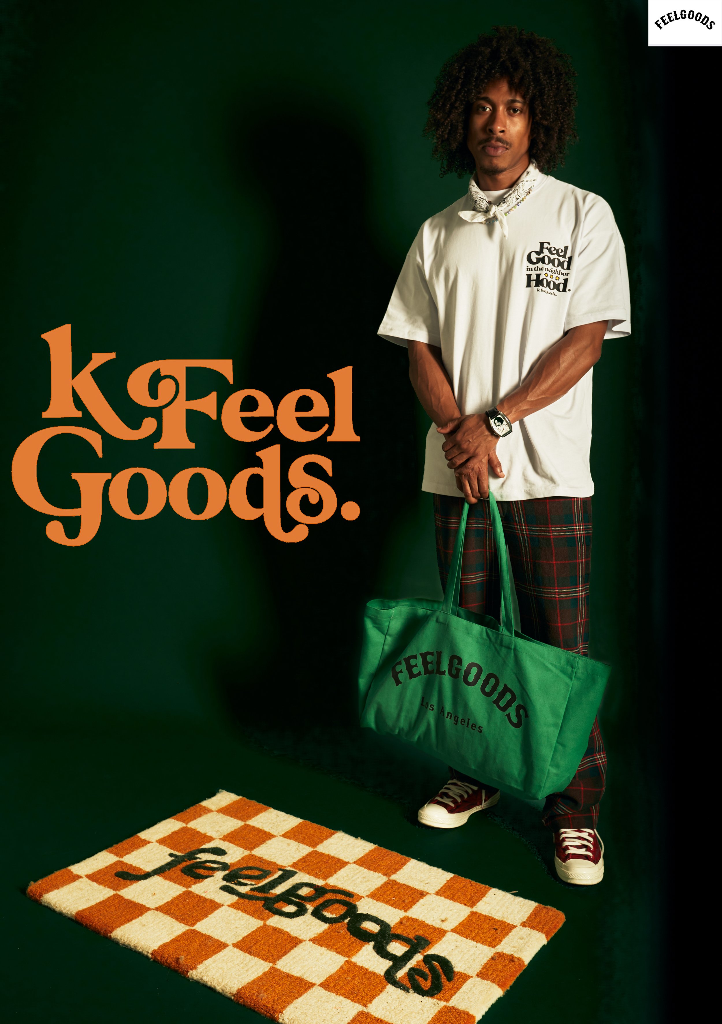 A man wearing a white Feel Goods t-shirt, plaid pants, and sneakers, standing on a checkered orange and white rug with the Feel Goods logo, holding a green tote bag with the Feel Goods logo, against a dark green background.