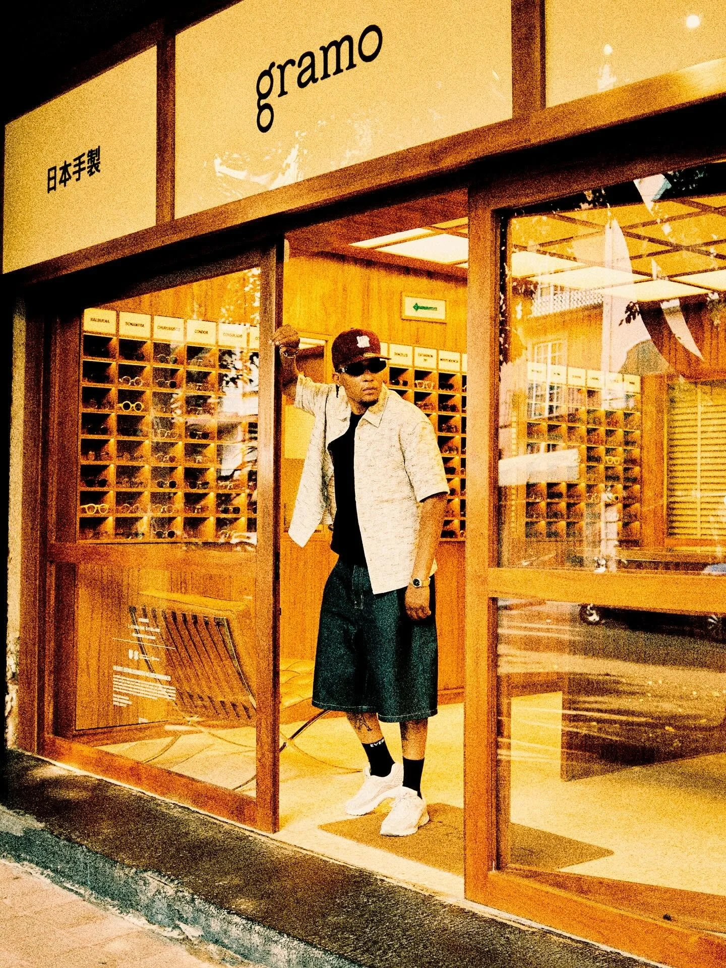 A man standing at the entrance of a wooden store with glass doors and windows, raising his right arm with a clenched fist and wearing sunglasses, a black cap, a beige short-sleeve shirt, black shorts, white sneakers, and black socks.