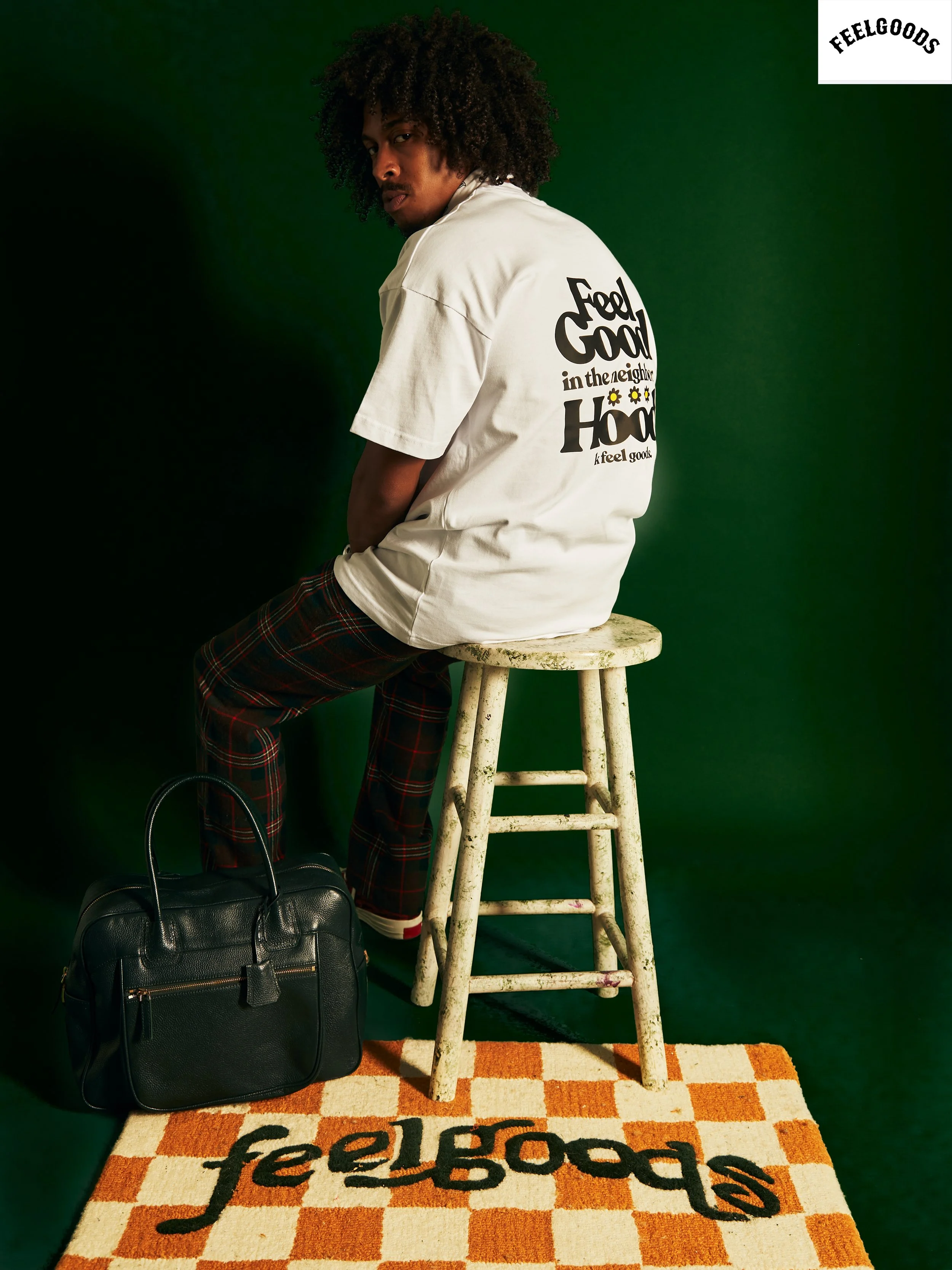 A young man with curly hair sitting on a white wooden stool against a green backdrop. He wears a white t-shirt with black text and illustrations on the back, plaid pants, and white sneakers. A black handbag rests on the floor near his feet, and there is a checkered rug with orange and cream colors featuring the word 'feelgoods' in black.