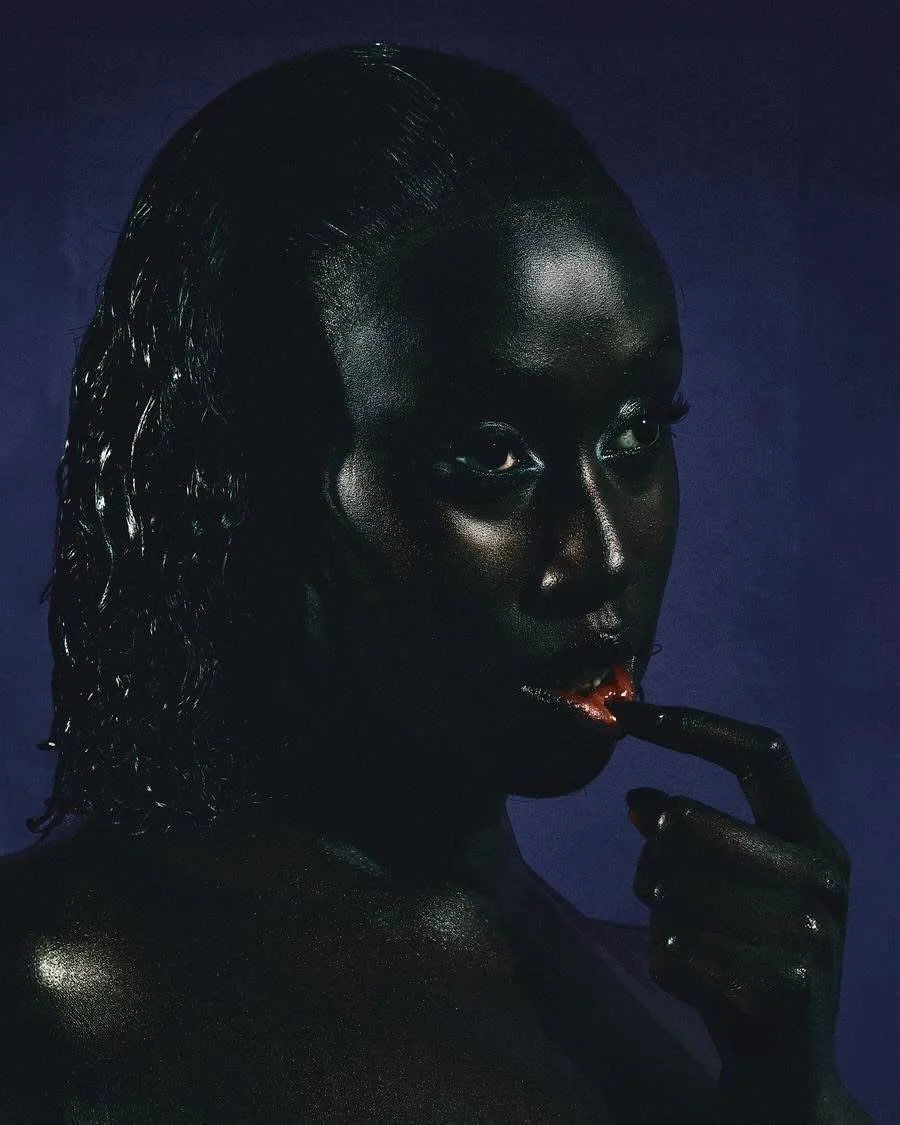 A woman with dark skin and wet hair touching her lips with her finger against a dark background.