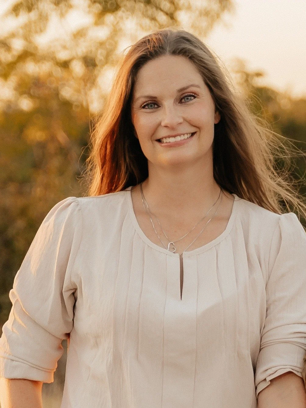 A smiling woman in a cream-colored blouse standing outdoors during sunset with blurred trees and warm lighting.