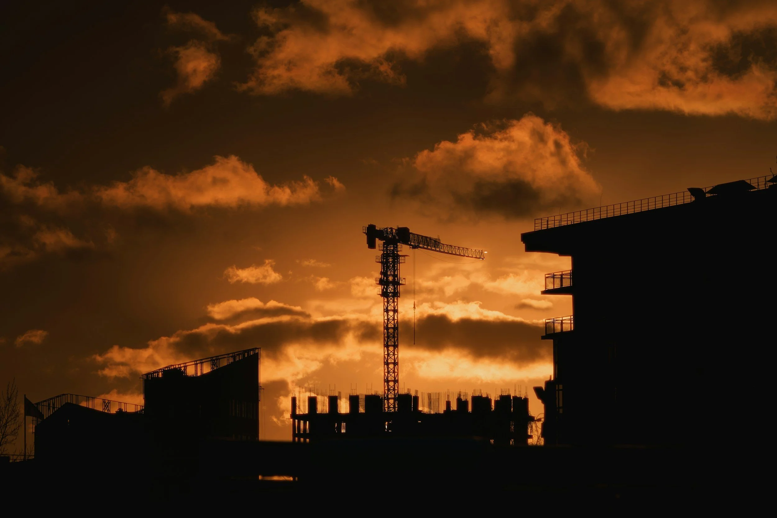 Silhouette of a construction crane and partly built buildings against a sunset sky with orange clouds.