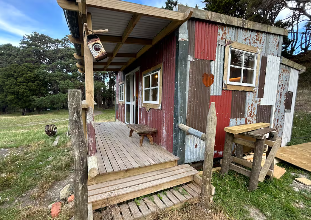 A small house made from rusty, multicolored metal sheets with a wooden porch and frame. The house has three windows with white frames, and a small decorative heart on the exterior wall. There is a wooden bench on the porch, and a rustic wooden railing with logs surrounds it. Nearby, there is a wooden table with an uneven surface, and various objects including a birdhouse hanging from the porch roof. In the background, there are trees and grassy open land under a partly cloudy sky.