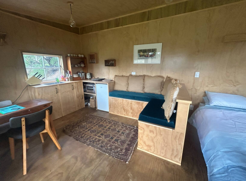 Small room with wood-paneled walls. Contains a bed with white bedding, a built-in seating area with cushions, a small kitchenette, a table with chairs, and a window with a view of trees.