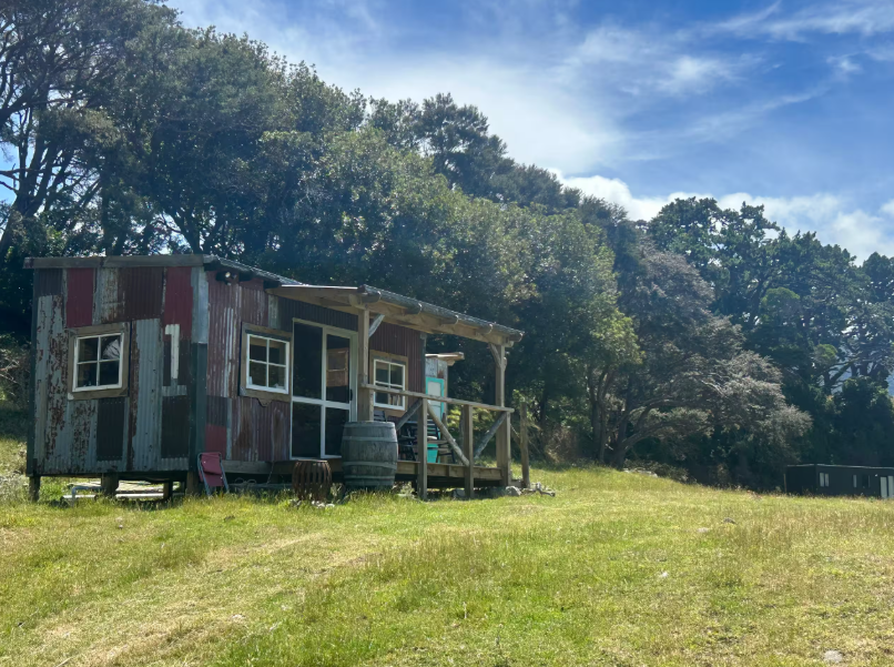A small rustic wooden cabin with a porch, situated on a grassy field with trees in the background.