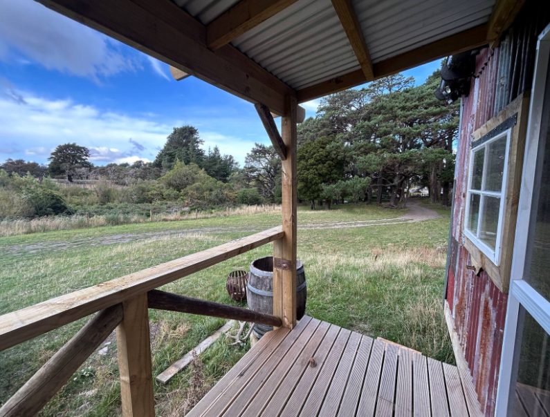 View from a rustic porch with wooden railing overlooking grassy field, trees, and a partly cloudy sky.