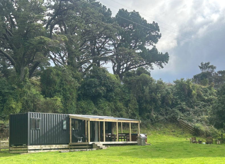 A small outdoor living space with a black shipping container as the base, a wooden deck with furniture, and a clear sky, surrounded by lush green trees and grass.