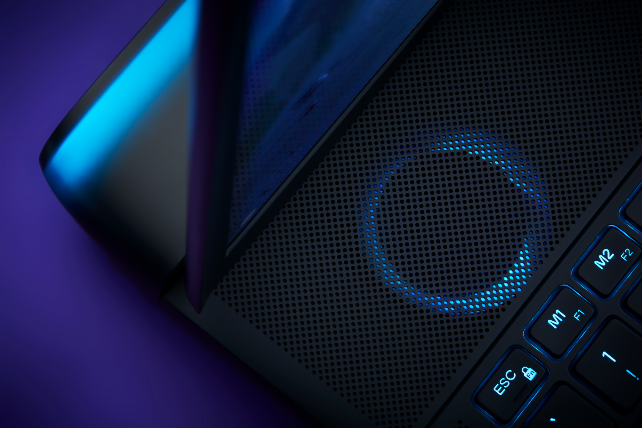 Close-up of a black laptop with a perforated surface, illuminated by blue LED lights and featuring labeled buttons such as M1, M2, F1, and F2.