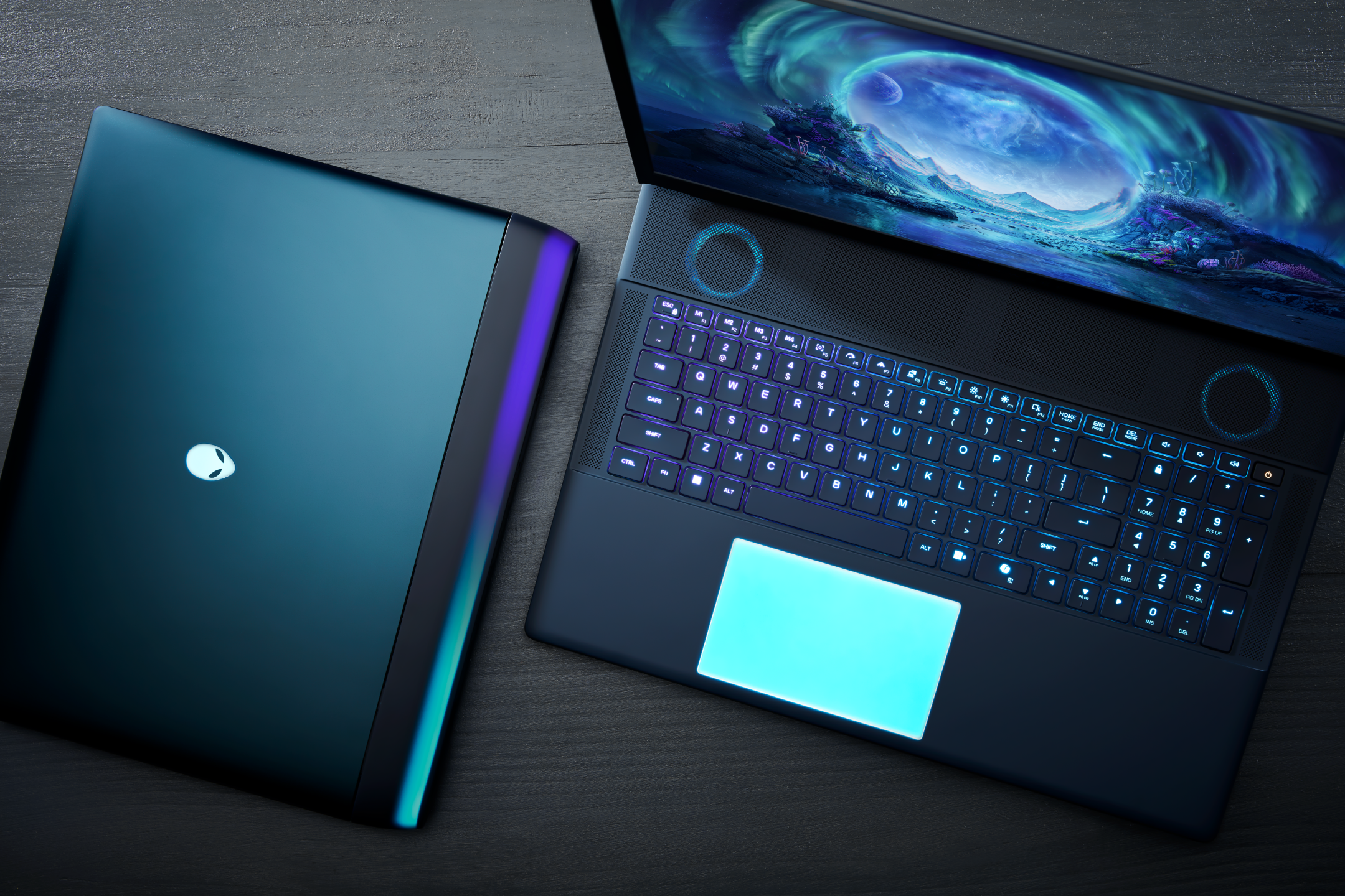 Top-down view of a gaming laptop with a colorful digital landscape on the screen, next to a closed Alienware laptop on a dark wooden surface.