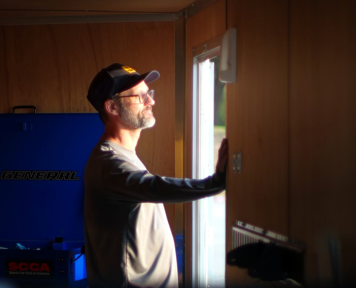 A man with glasses and a beard wearing a black cap and a jacket, standing inside a wooden room near a window, looking outside with sunlight on his face.
