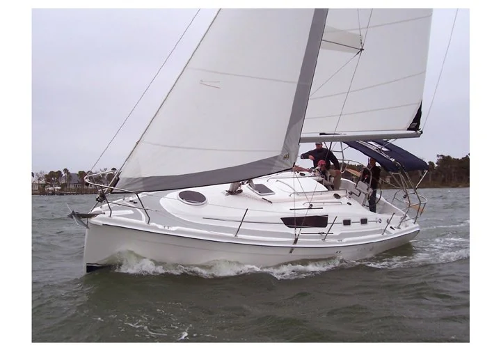 A sailboat with white sails cruising on the water, with two people on deck adjusting equipment, and a shoreline with trees and buildings in the background.