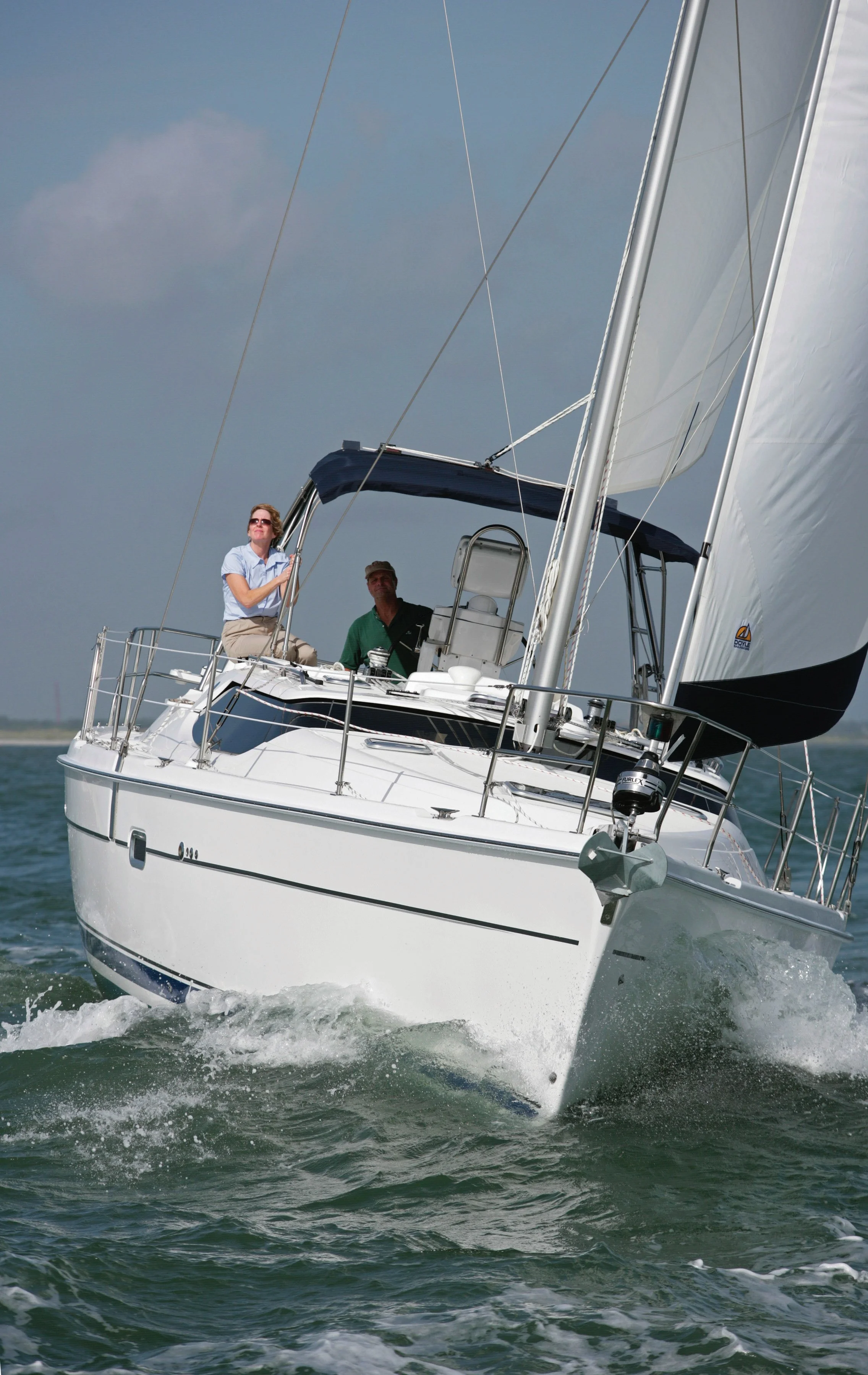 A white sailboat with two people on board sailing on the water under a blue sky with some clouds.