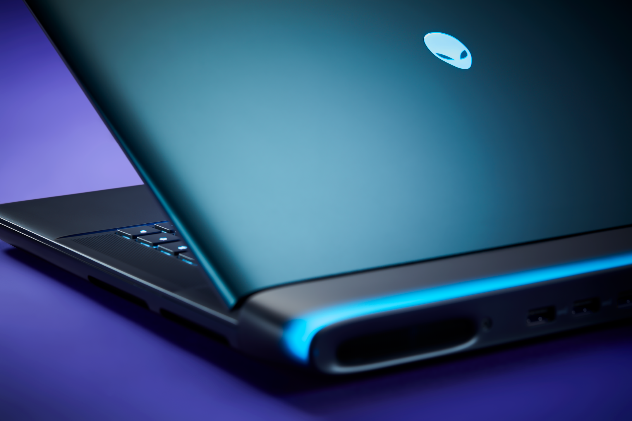 Close-up of a laptop with a glowing alienhead logo on the lid, with part of the keyboard visible and illuminated keys, set against a dark background with blue and purple lighting accents.