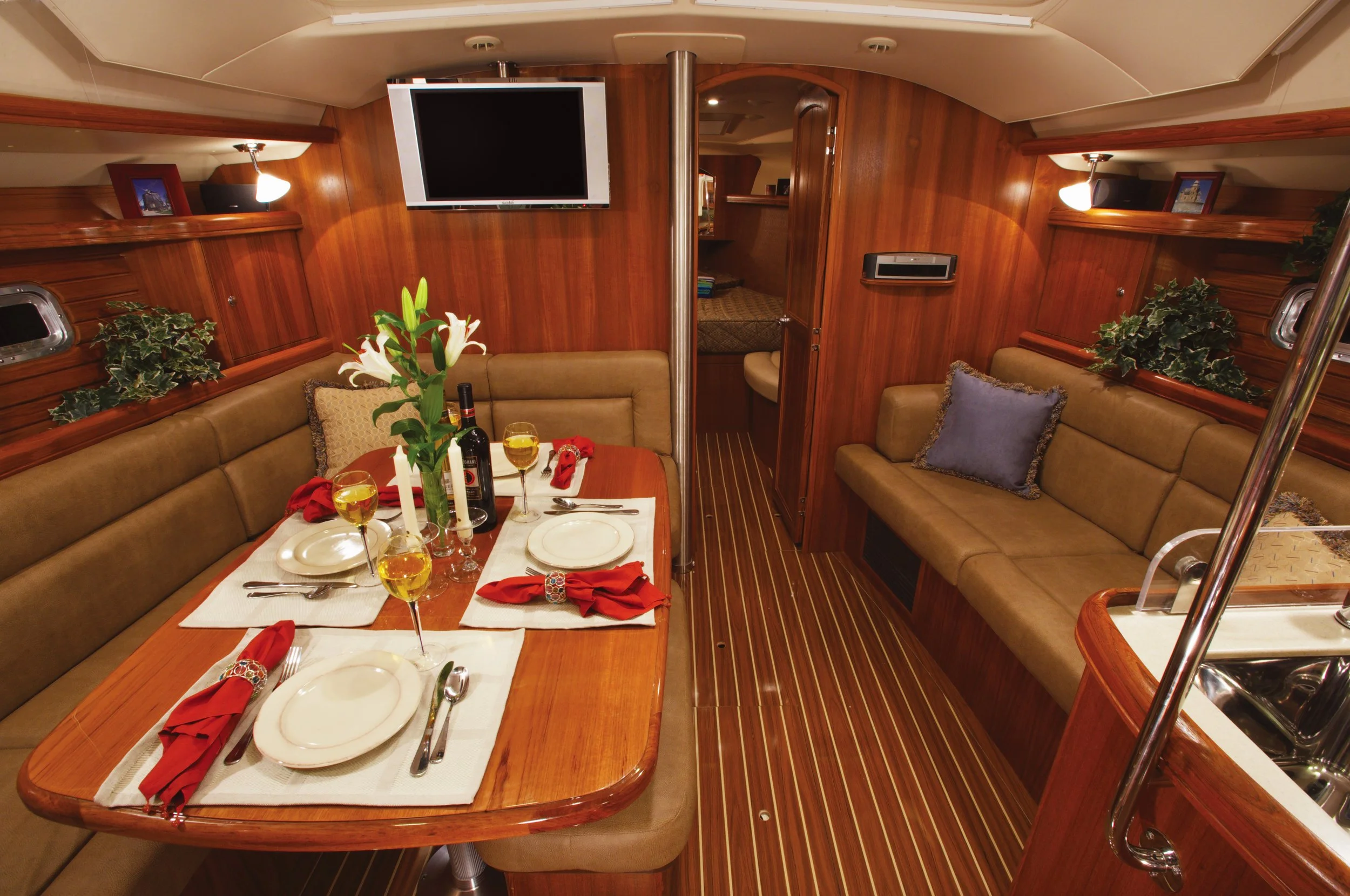 Interior of a yacht with a wooden dining area, set for a meal with plates, red napkins, wine glasses, and a centerpiece with white lilies. There are two built-in sofas with cushions, a mounted flat-screen TV, small framed pictures, plants, and a small window.