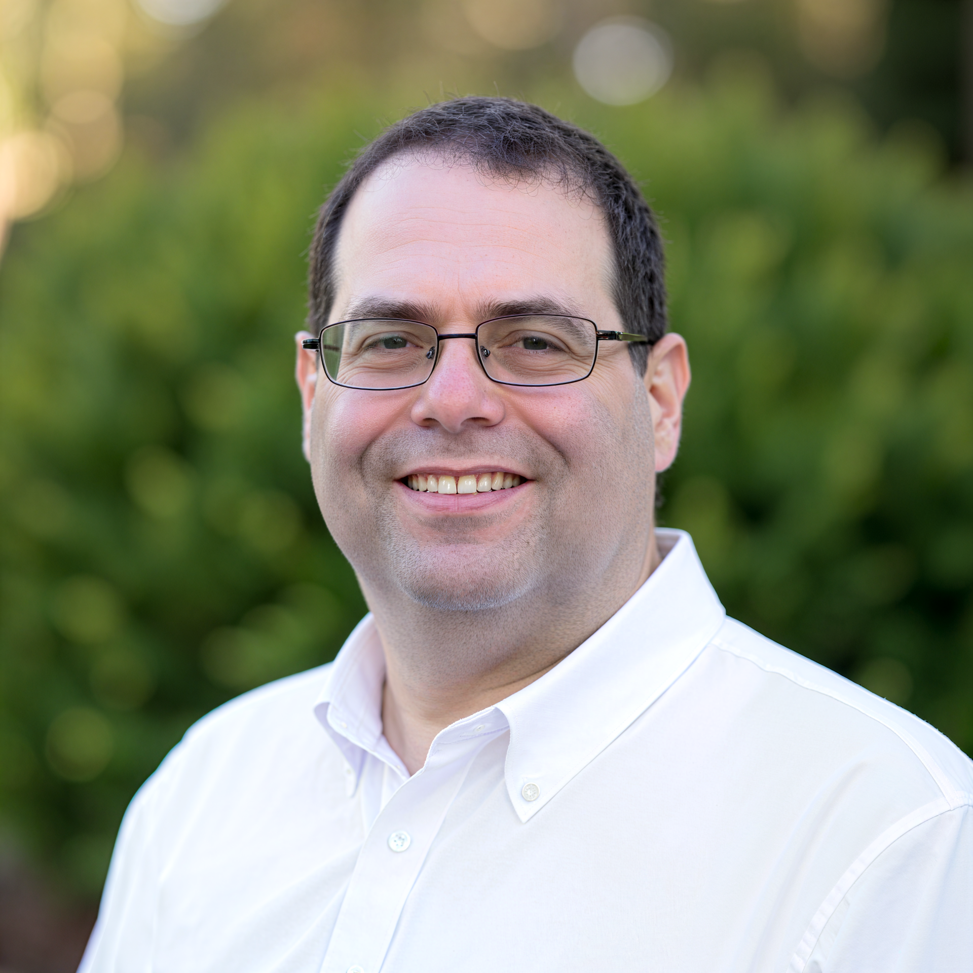 Neal A. Levene is wearing a white shirt, outdoors with a blurred green background.
