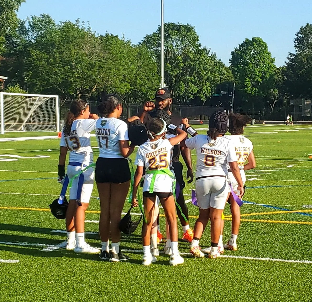 A youth football team gathered around their coaching staff on a football field during practice, with trees and goalposts in the background.
