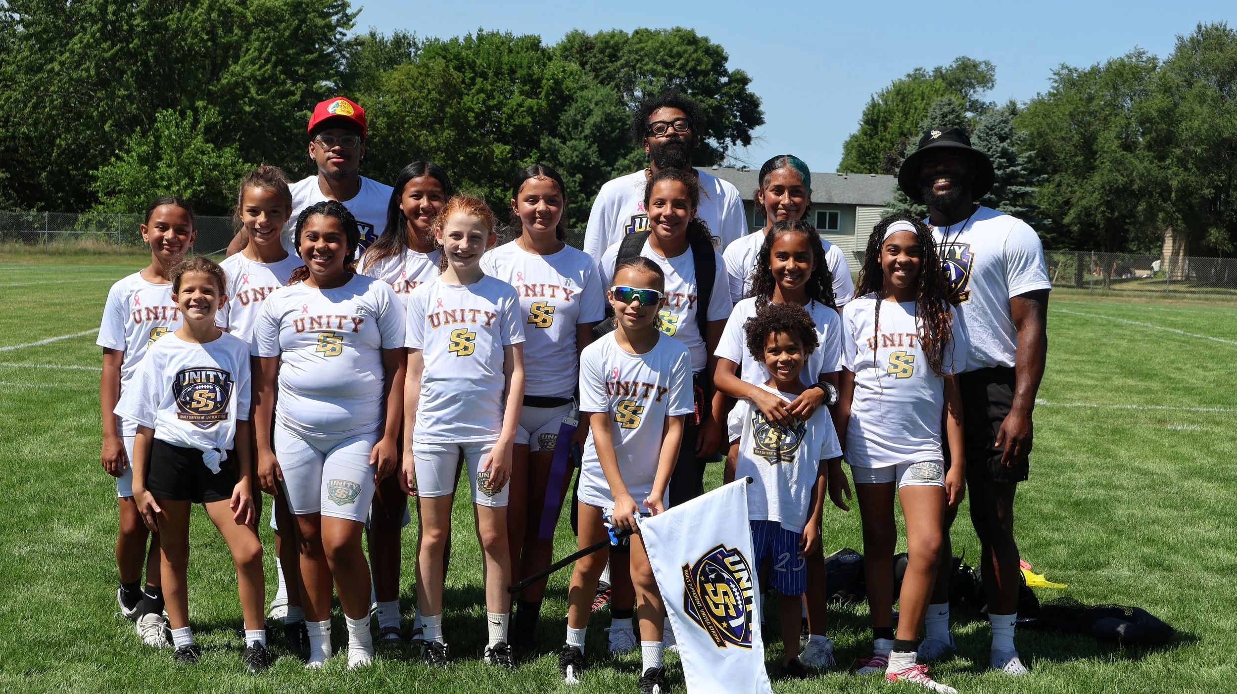 Group of young girls and two adult men on a grassy field, wearing white sports shirts with 'Unity' and a logo, posing for a photo after a sports event, with trees and houses in the background under a clear blue sky.
