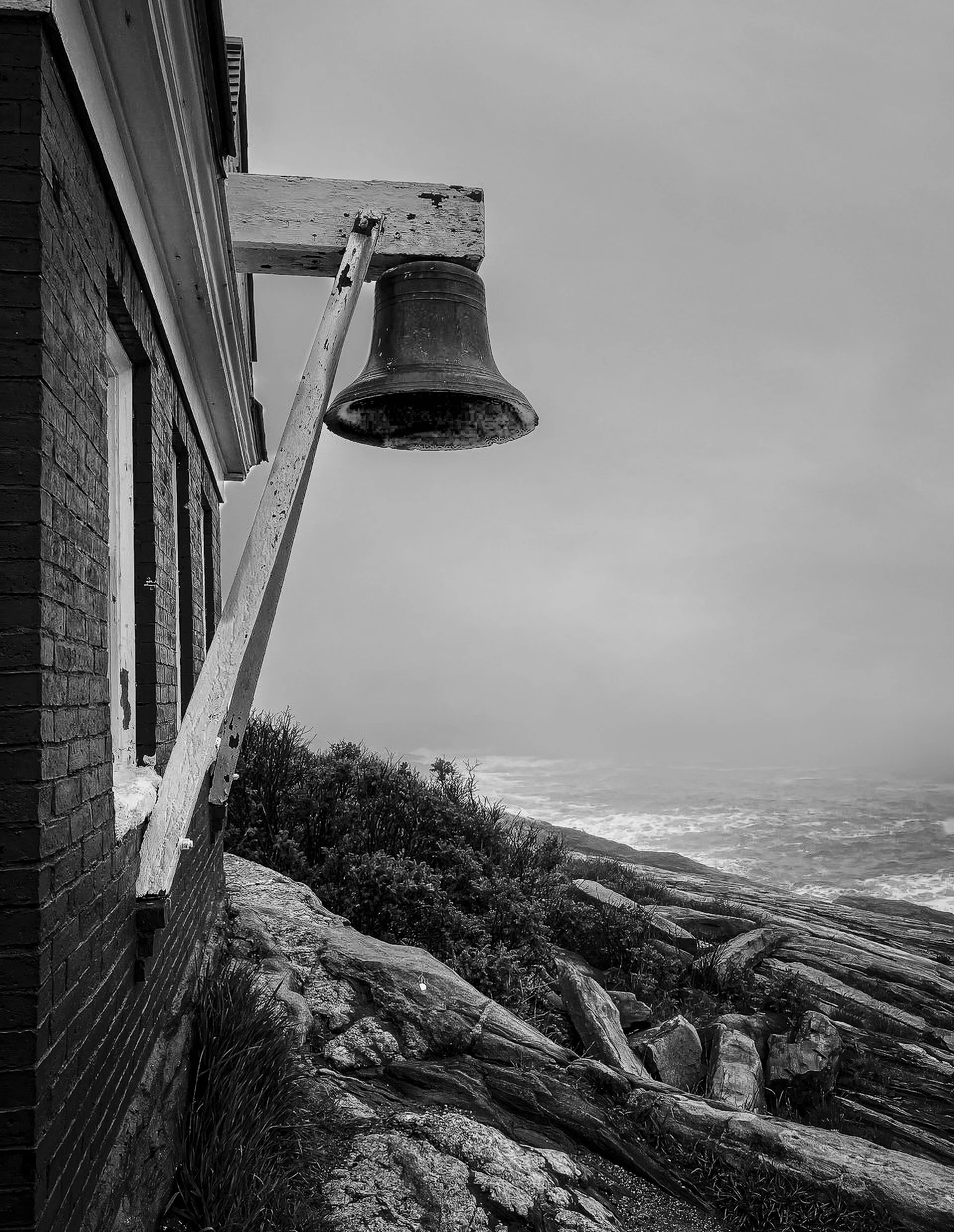 Black and white photo of a building's brick wall with a window on the left, and a clock at the top. A wooden ladder leans against the wall, leading to a wooden platform with a large bell hanging from it. The platform is supported by a wooden beam. The view extends to a rocky coastline with sparse vegetation and the ocean in the background.