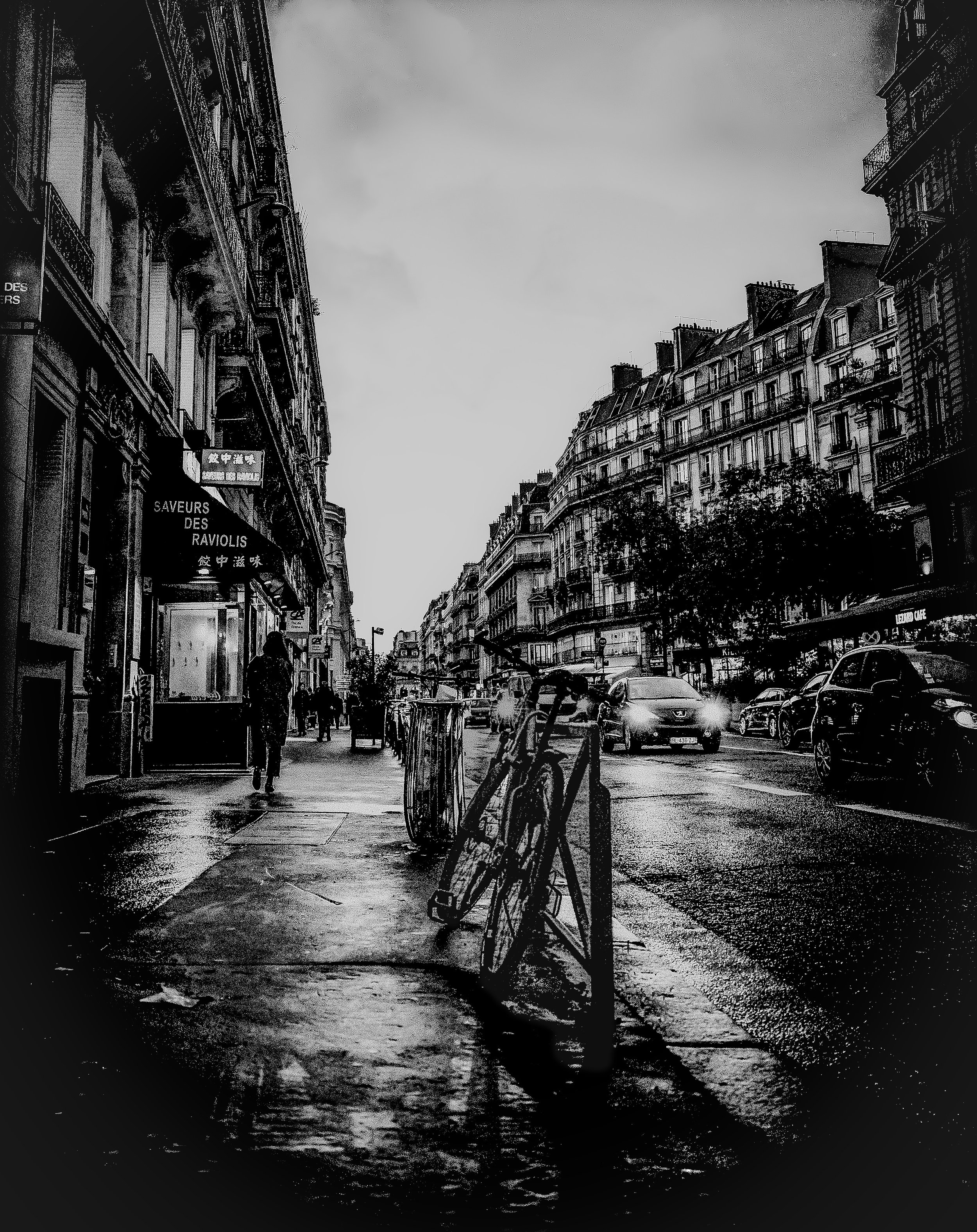 A black and white photo of a city street scene. On the left side, there are storefronts with signs, including one that reads 'Saveurs Des Raviolis'. The right side has residential and commercial buildings with balconies. Pedestrians walk along the wet sidewalk, and there are several cars on the street, some with headlights on, indicating it might be dusk or rainy.
