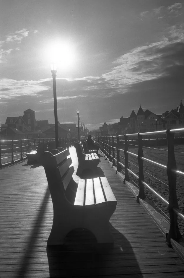Black and white photo of a boardwalk with empty benches, a person sitting on one bench, lamp posts, and houses in the distance, with a bright sun and partly cloudy sky overhead.