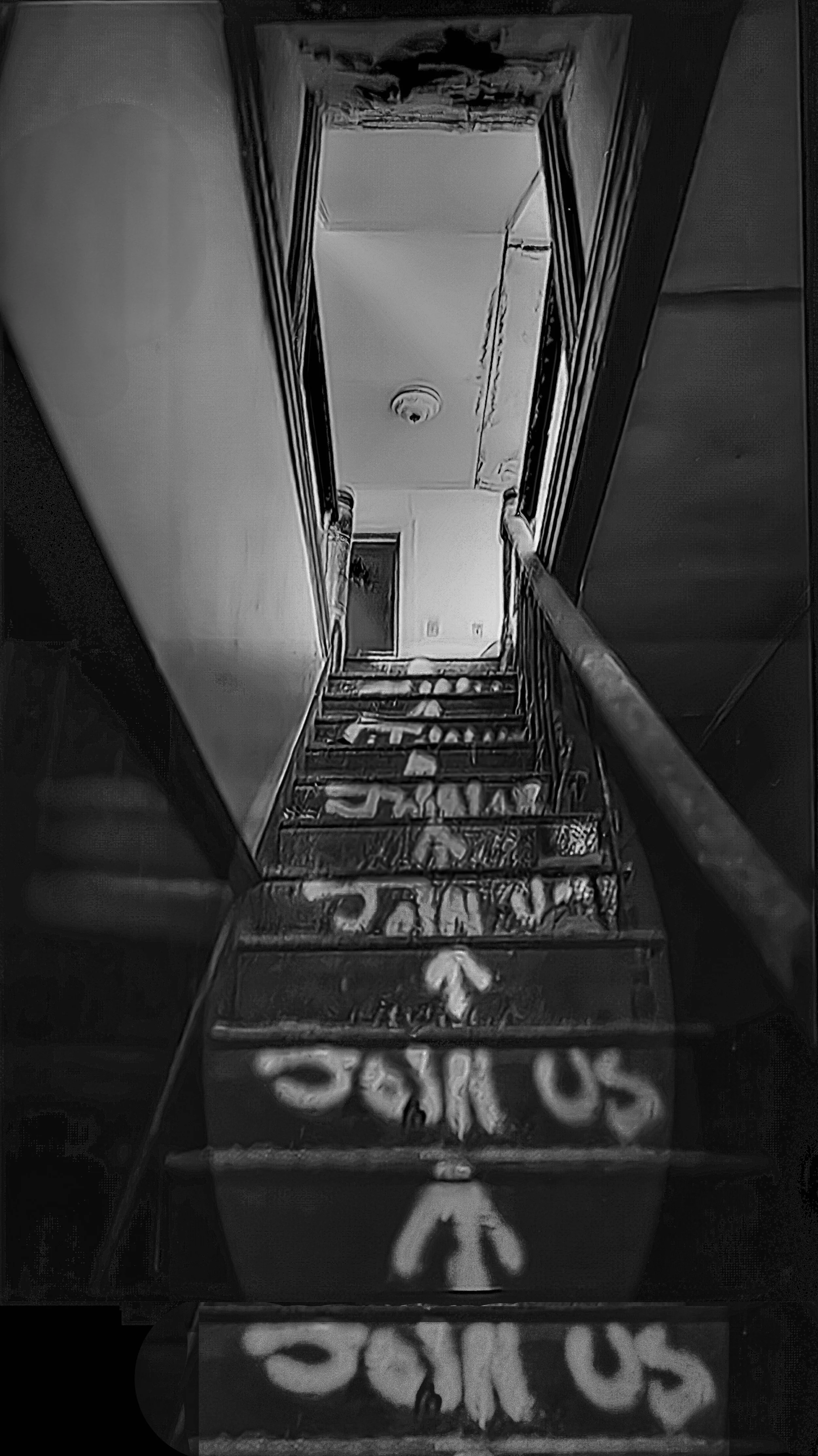 Black and white photo of a staircase with a handrail, leading to a door at the top, with a fire alarm on the ceiling and patterned carpet on the steps.