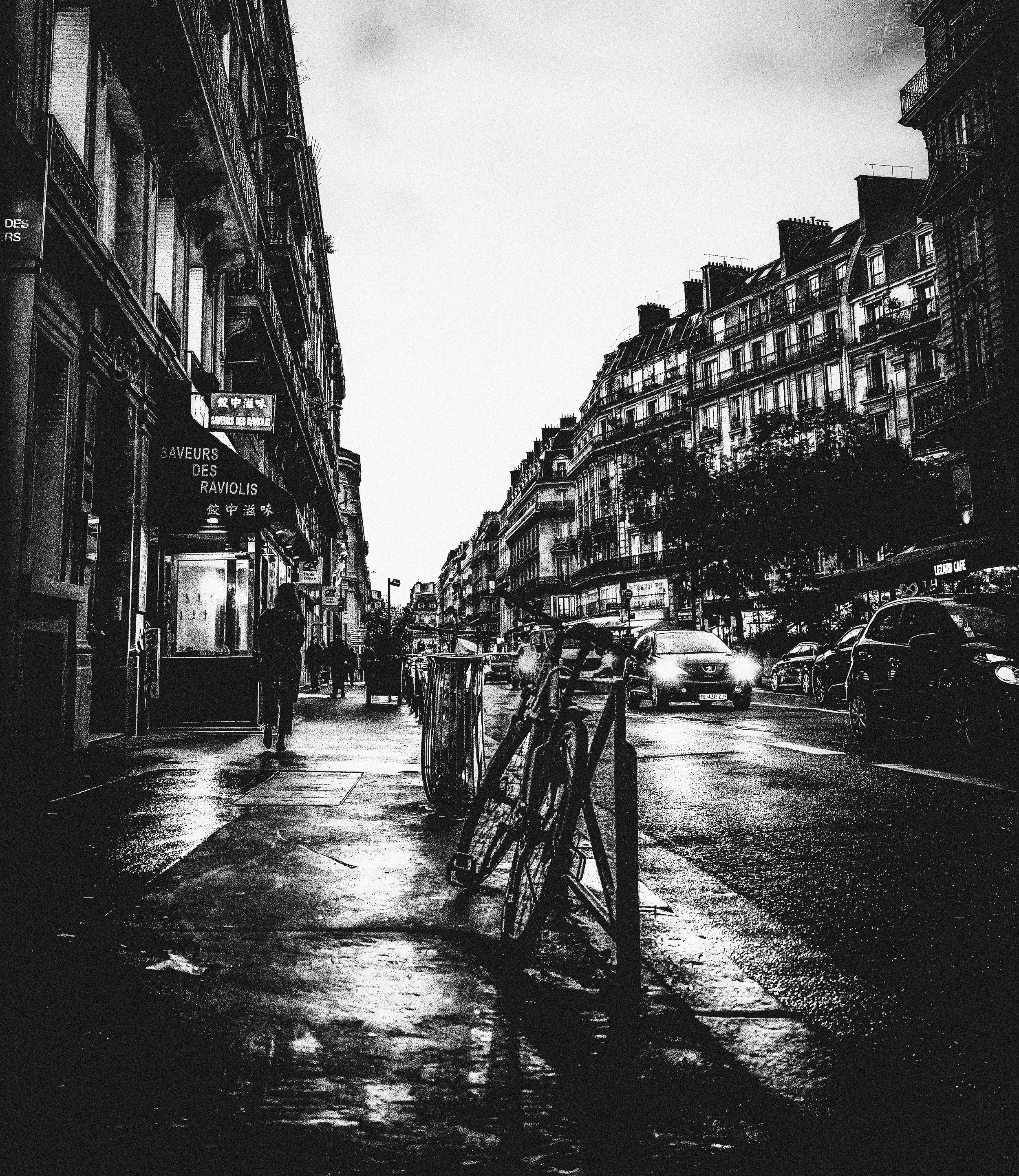 A black-and-white photo of a city street during sunset or rain, with Parisian-style buildings on either side, cars parked along the street, and a few pedestrians walking. There are outdoor cafes and signs in French.