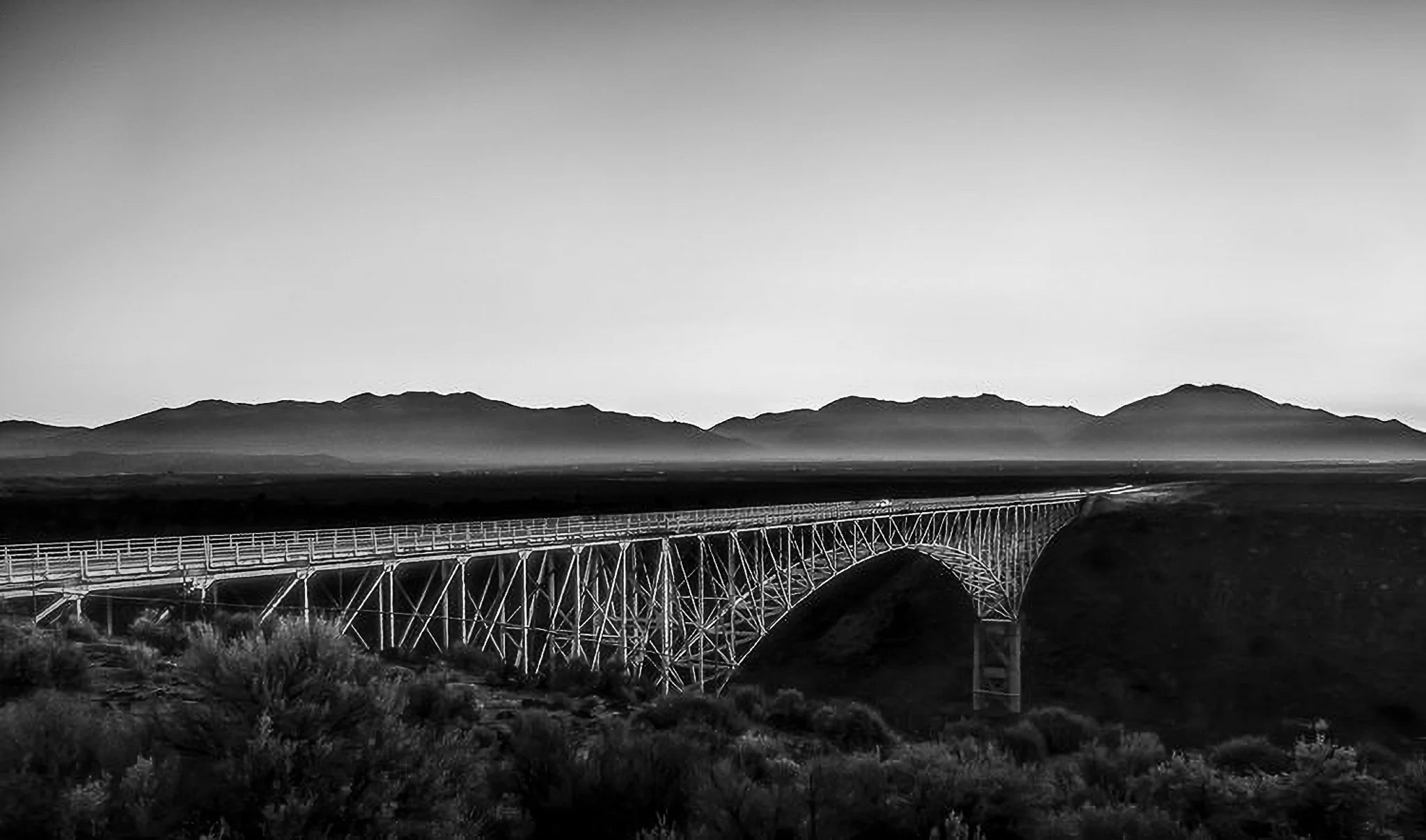 Black and white photo of a bridge spanning a valley with mountain range in the background.