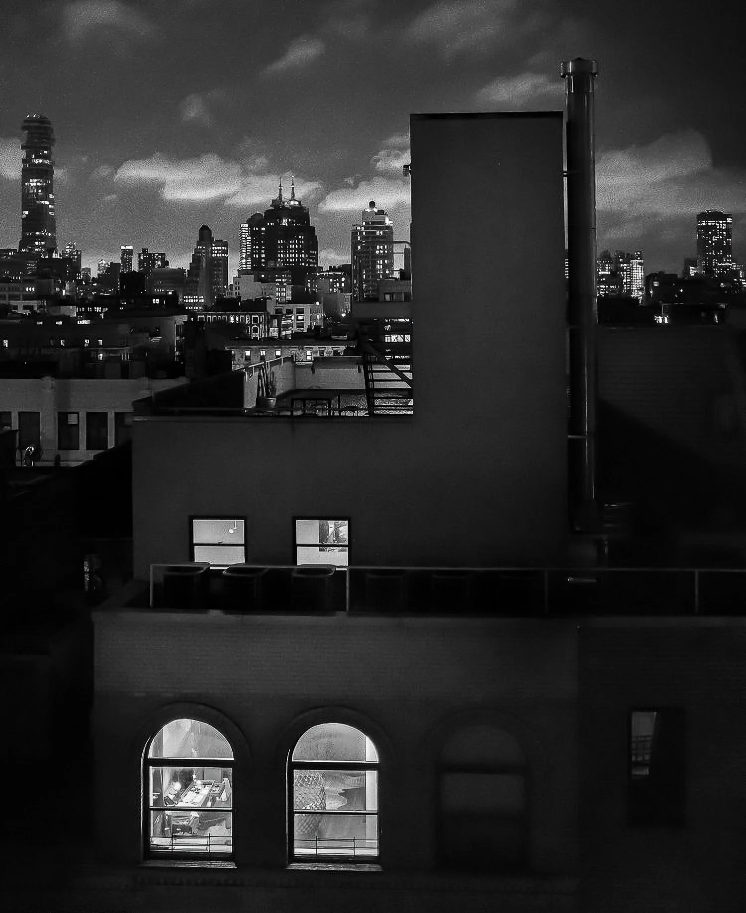 Nighttime cityscape with illuminated buildings, seen from a rooftop with windows and a small terrace.