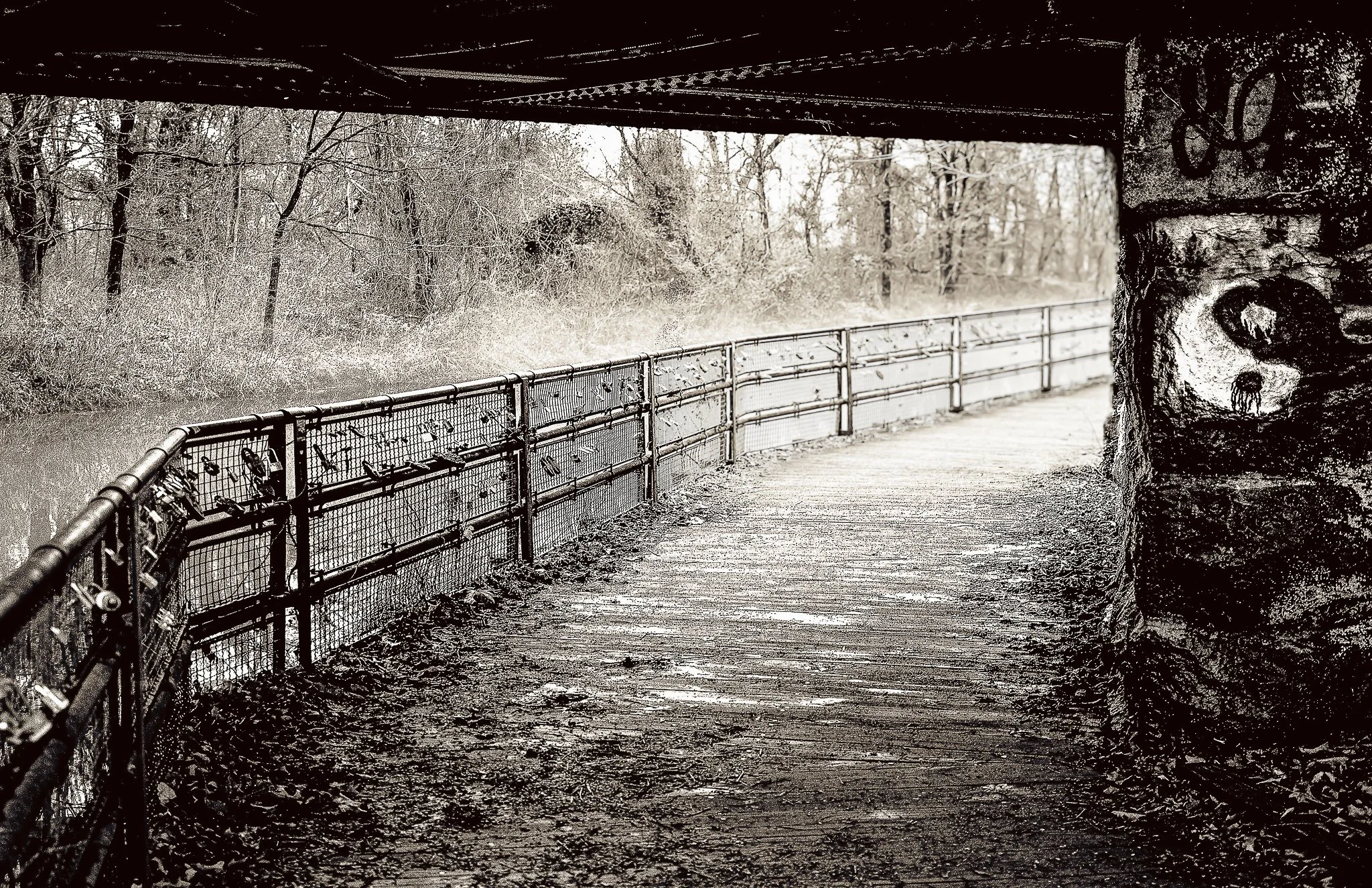 A snow-covered pathway under a bridge with a fence running alongside, trees in the background, and snowfall in a winter scene.