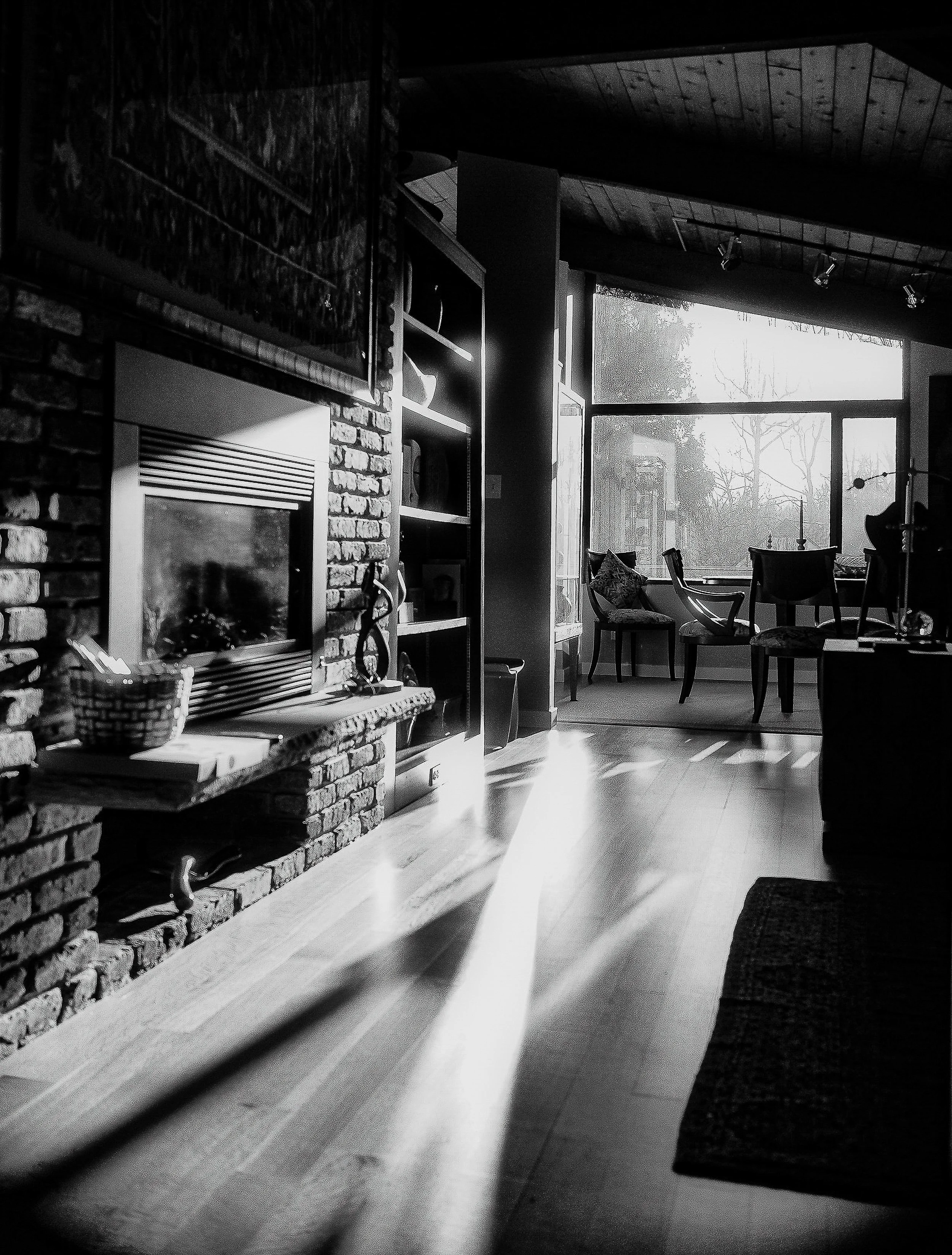 Black and white photo of a cozy living room with a large window, chairs, a sofa, a brick fireplace, and indoor plants, illuminated by natural light.