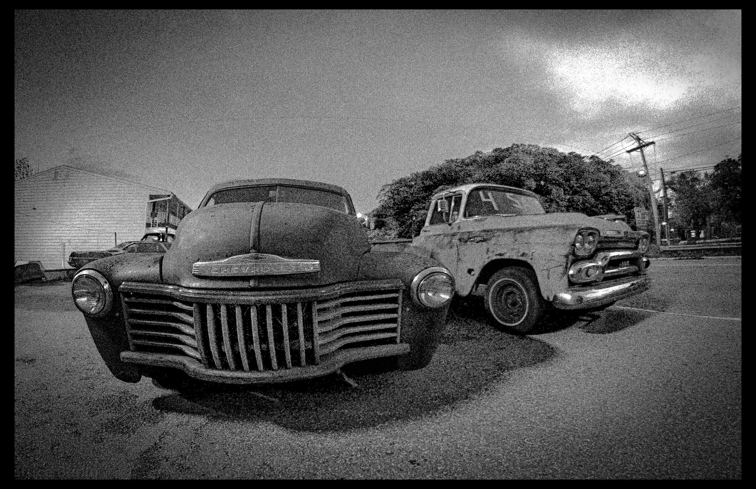 Black and white photo of vintage cars parked outdoors at night, with a large tree, utility poles, and buildings in the background.