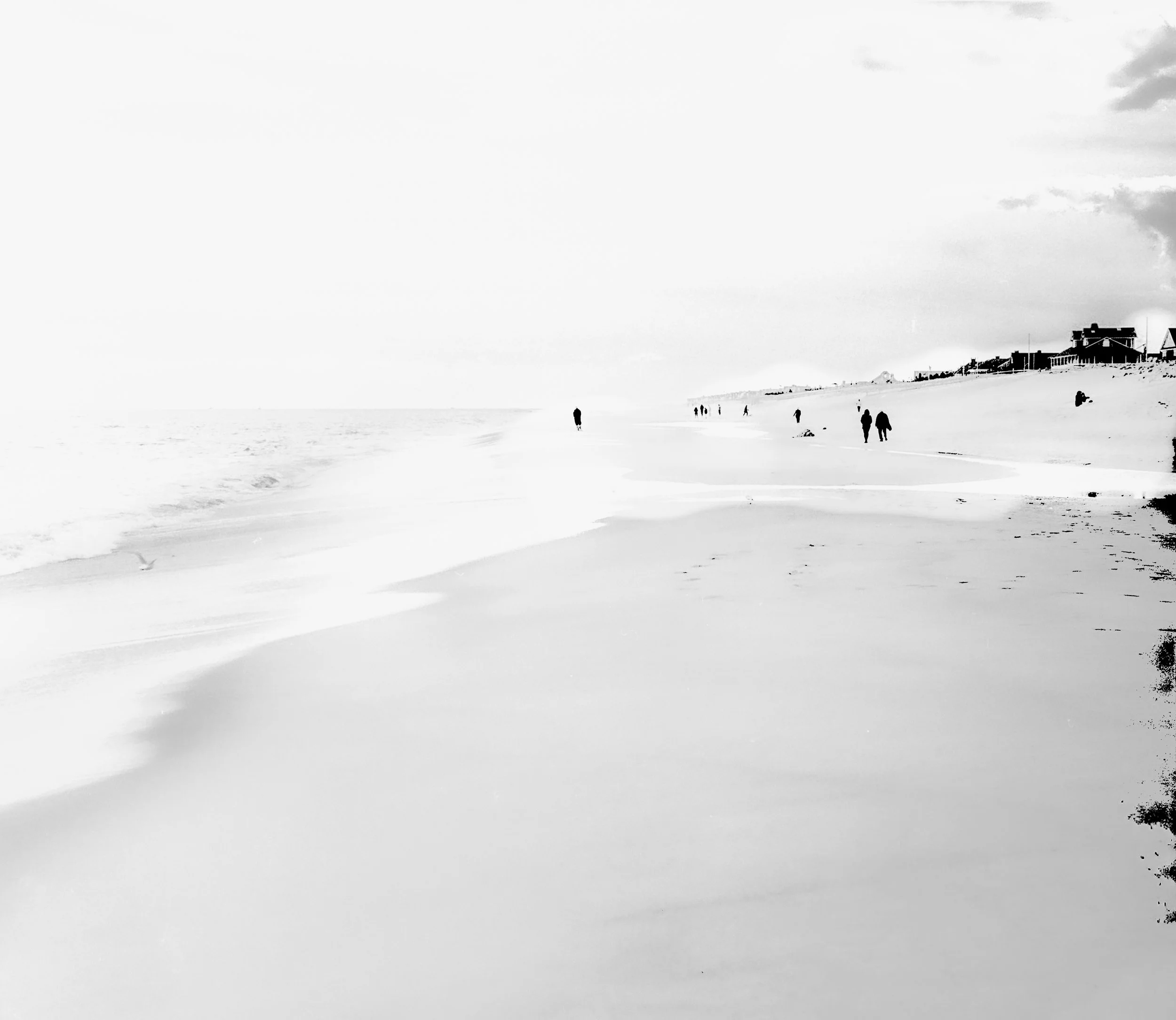 A black and white photo of a beach with several people walking along the shore and houses on a hill in the distance under a cloudy sky.