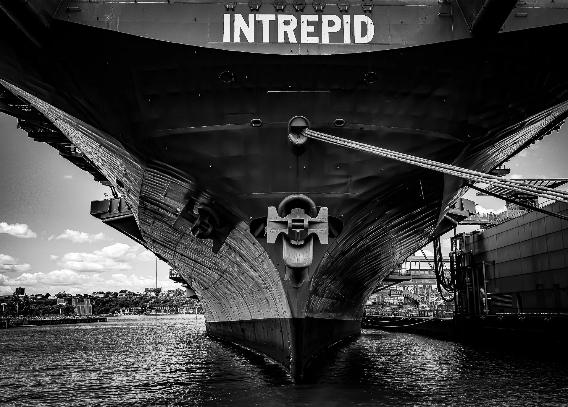 Close-up view of the bow of a large, modern battleship named Intrepid, docked at a harbor with a city skyline in the background. The image is in black and white, showing the ship's hull and anchor details from a low angle.