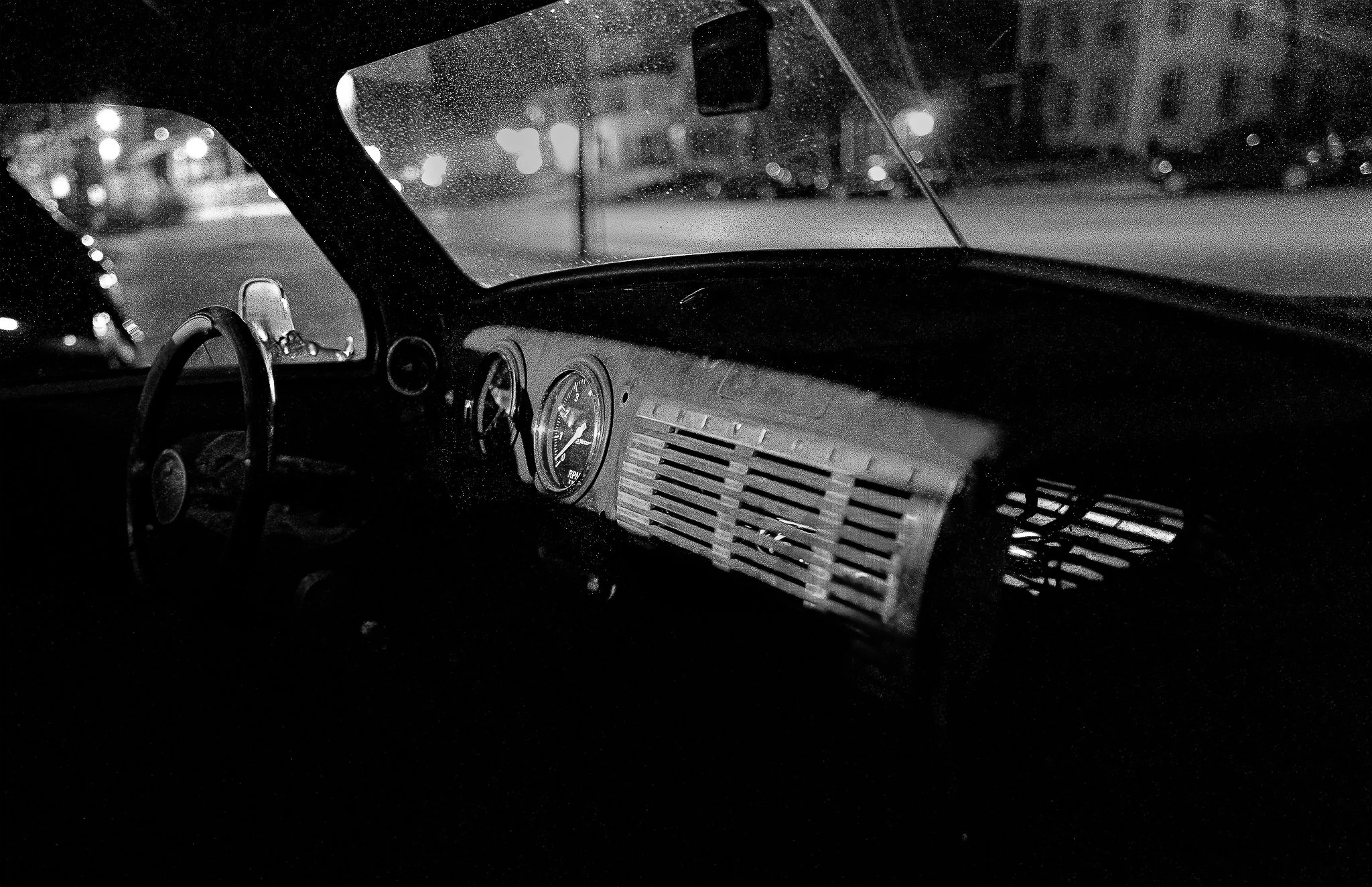Nighttime view of a car's interior with the dashboard and steering wheel visible, showing rain droplets on the windshield and blurred streetlights outside.