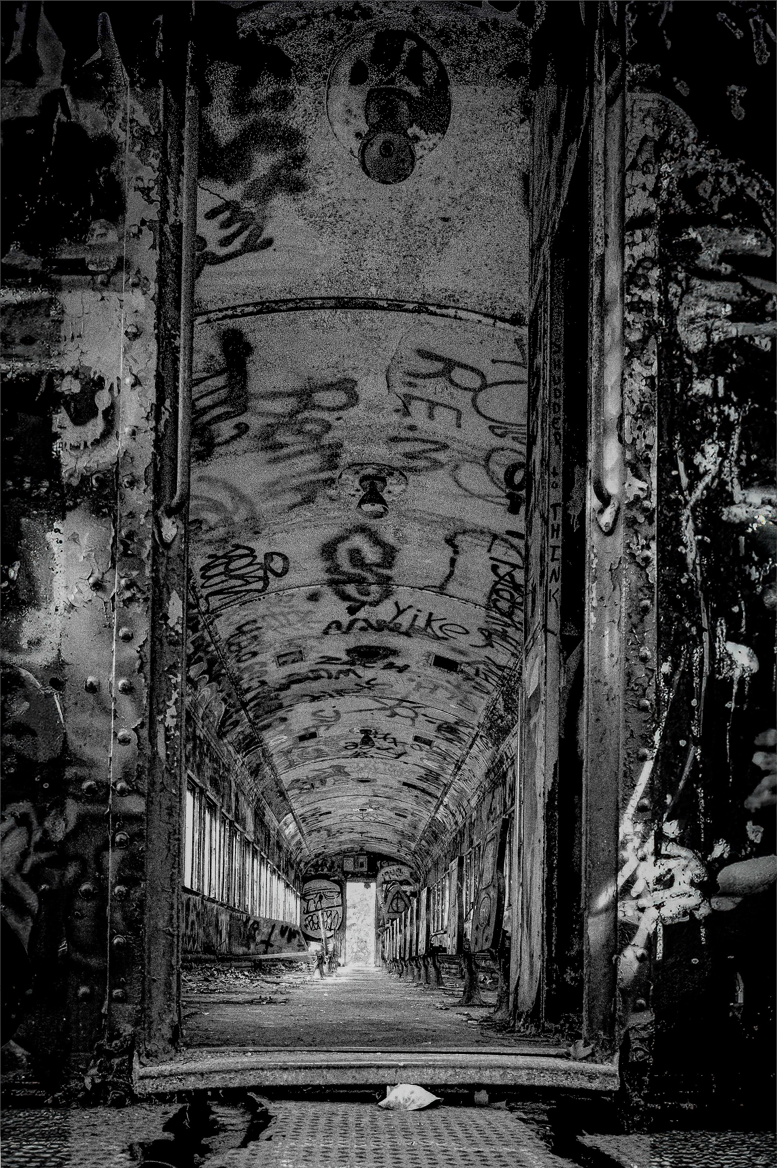 A long abandoned graffiti-covered corridor with a high arched ceiling and broken windows, leading to a lighted exit at the far end, with scattered debris and graffiti on the walls.