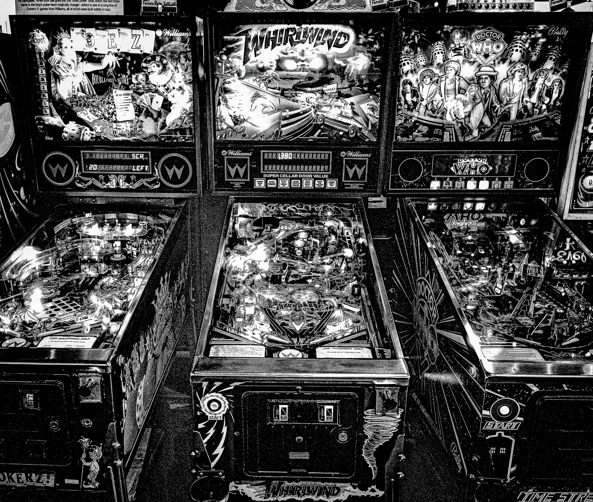 Black and white photo of several pinball machines, primarily featuring arcade-themed designs, with visible artwork and controls.