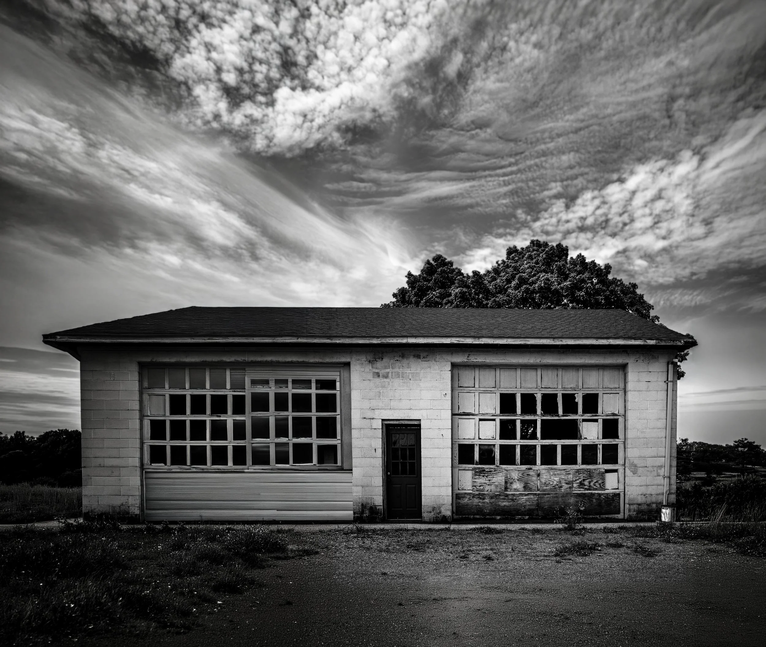 A black and white photograph of a house with a garage, a door, and large front windows, with a large tree and a dramatic sky with high cloud formations in the background.