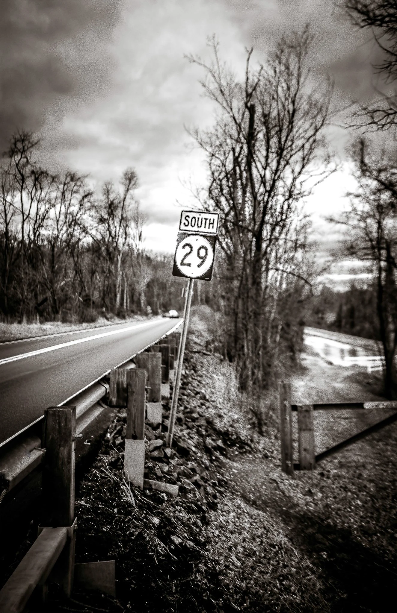Black and white photo of a rural highway with a southbound route 29 sign leaning, weathered guardrail, leafless trees, and a cloudy sky.